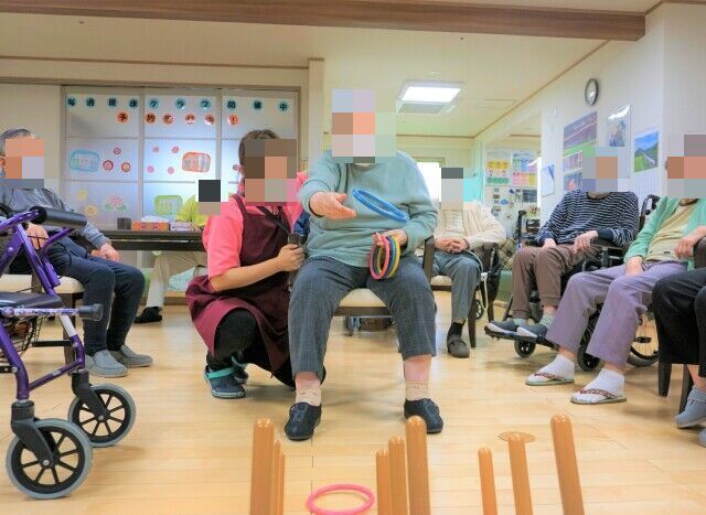 真ん中に赤いネットのついたカゴが置かれている。その周りを椅子に座った高齢者が囲んでいる。