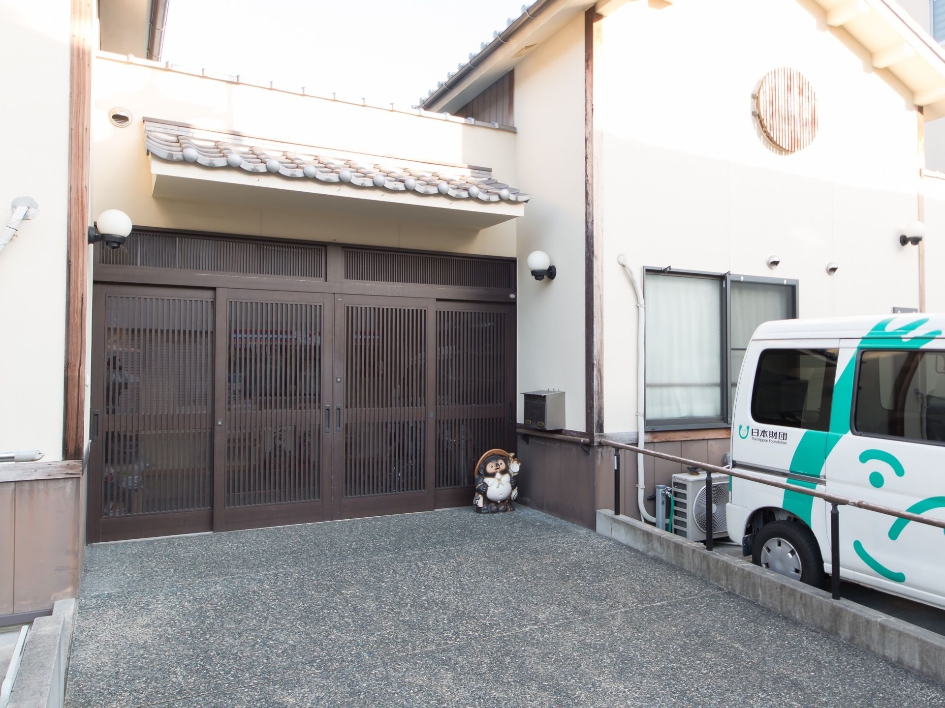 玄関には茶色い格子のドアが設置されていて、上には瓦の屋根がある。両サイドの壁に丸いライトが取りつけられている。