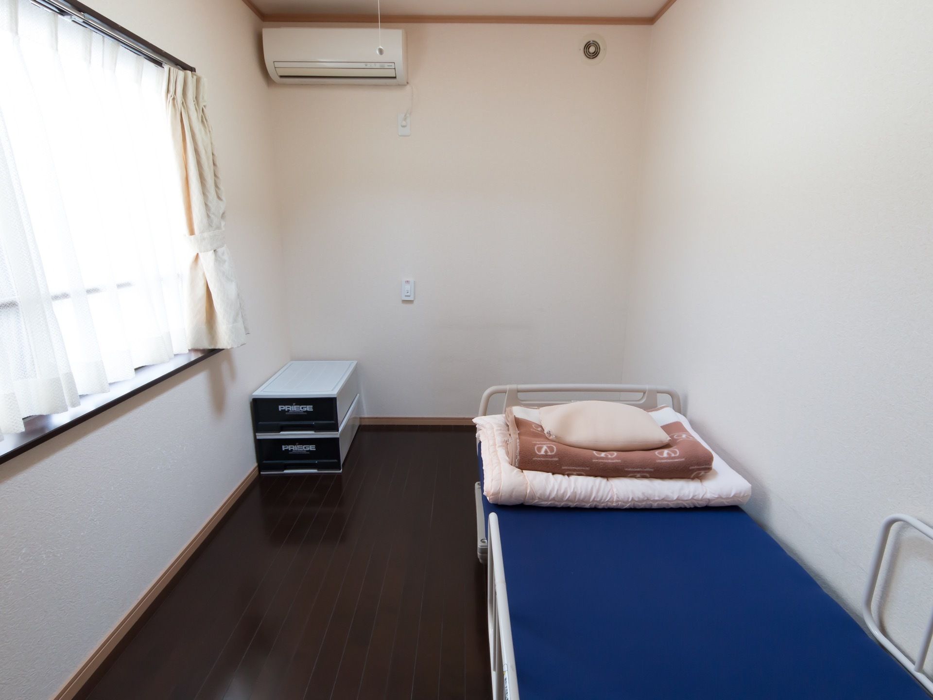 壁際に青いベッドが設置されていて、上にたたんだ布団と枕が乗せられている。部屋の隅に収納ケースが２つ重ねて置かれている。