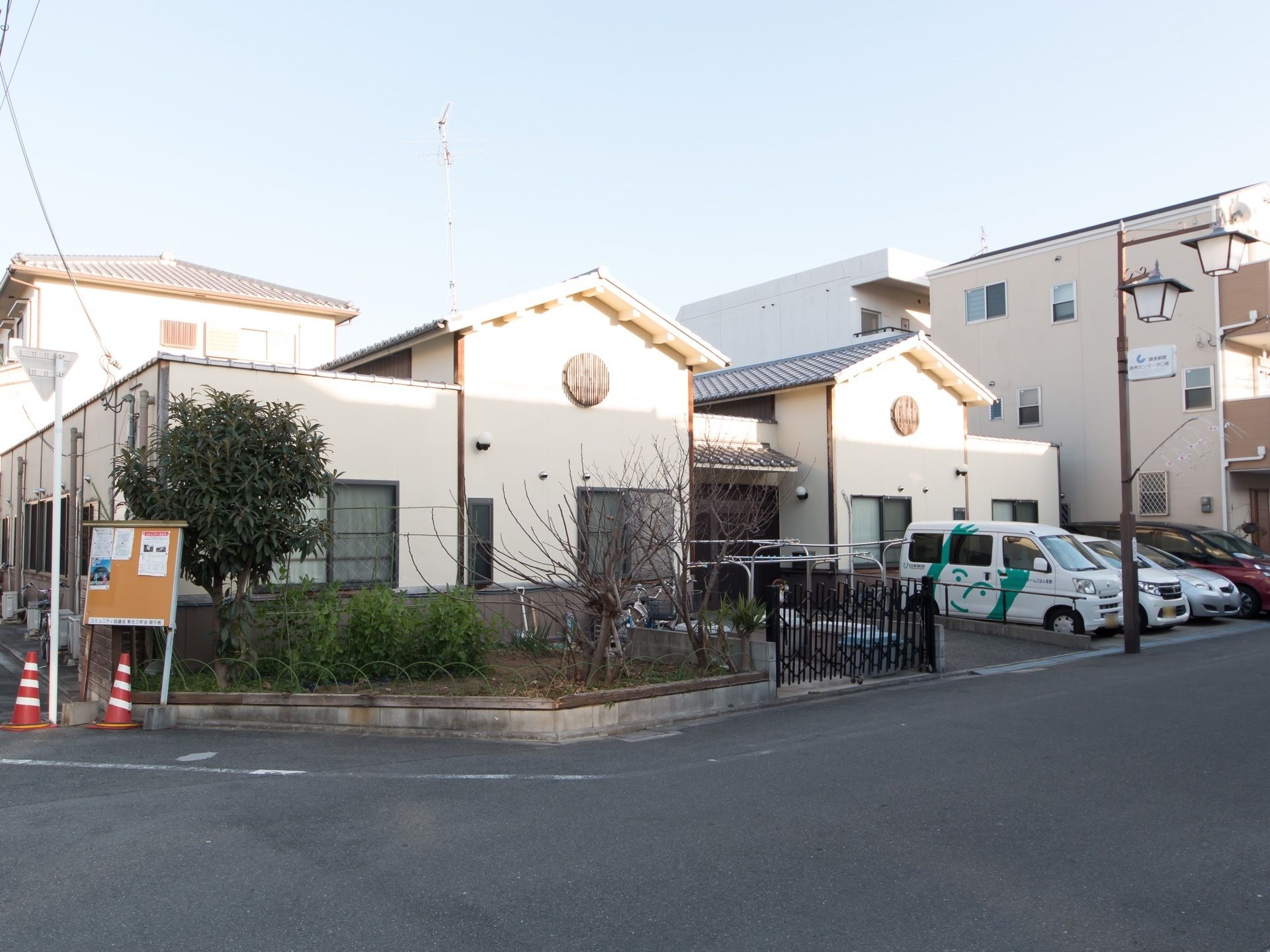 建物の前には庭があり、木や植物が植えられているのが見える。その前のコーナー部分に掲示板が設置されている。