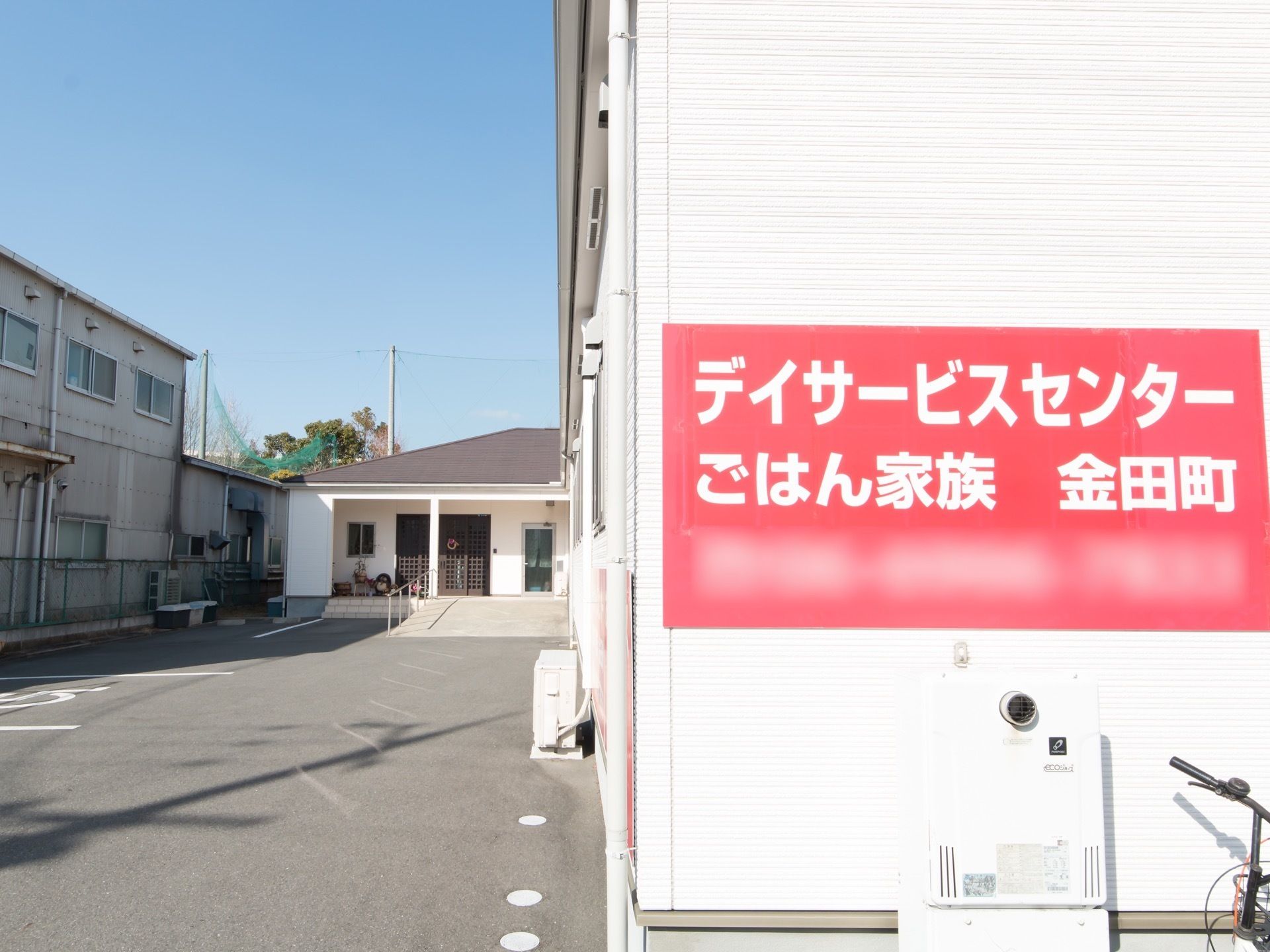 平屋建ての建物がある。玄関先の広い敷地の地面は舗装され、駐車場になっている。入り口には施設名が書かれた赤い看板が掛かっている。
