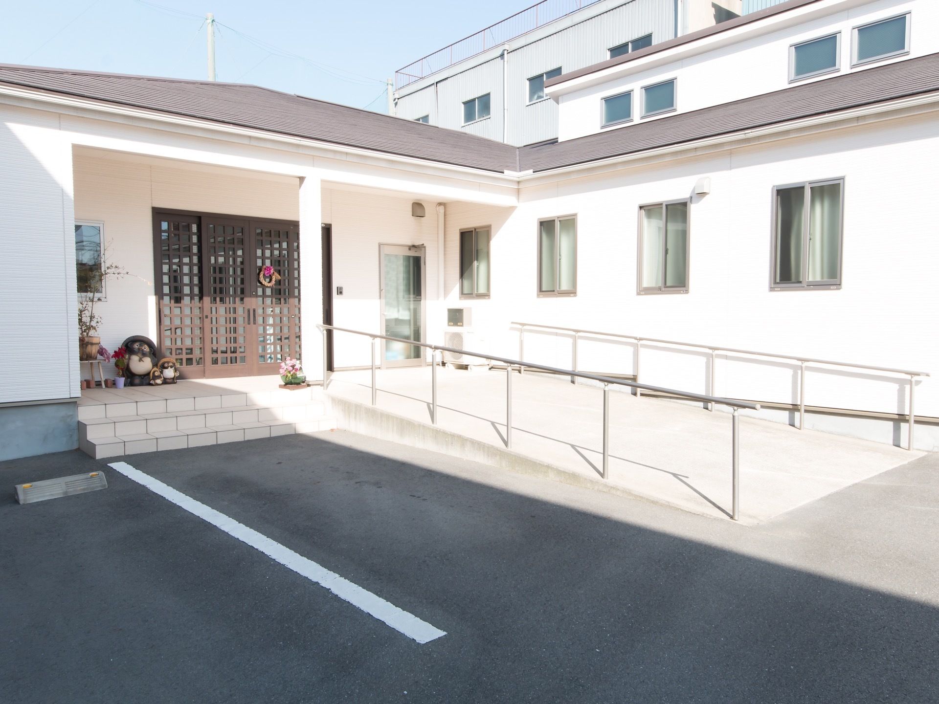 平屋建てのL字型の建物がある。玄関前は広く、階段が３段と大きなスロープ、手すりがある。脇には信楽焼きの置物と植木鉢が置かれている。