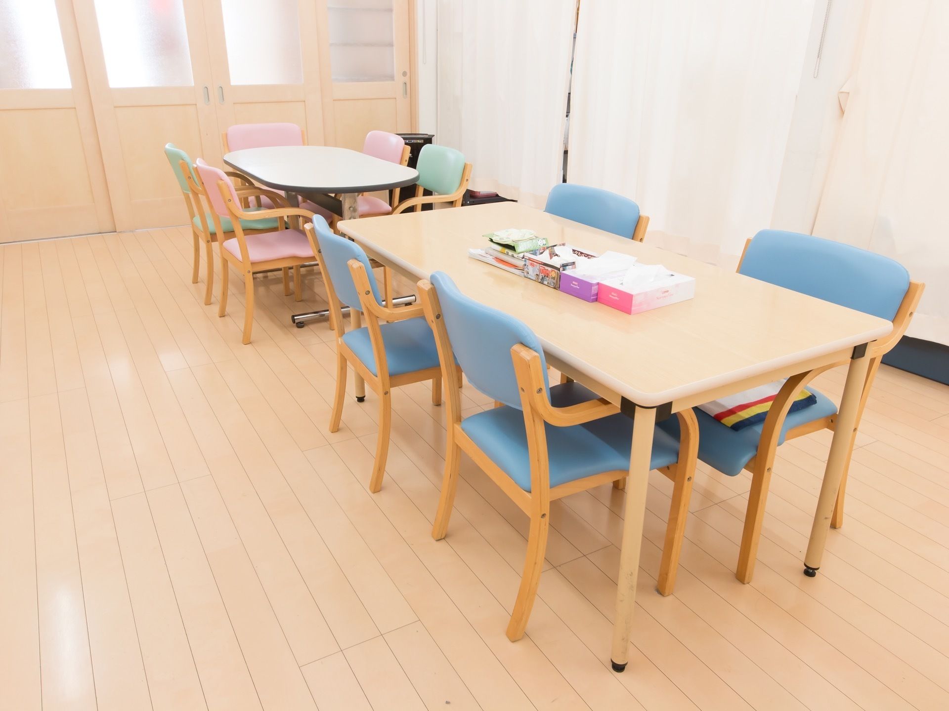 明るいベージュの床とドアのある食堂。大きなテーブルと椅子が並べられている。テーブルの上にはティッシュ箱が置かれている。