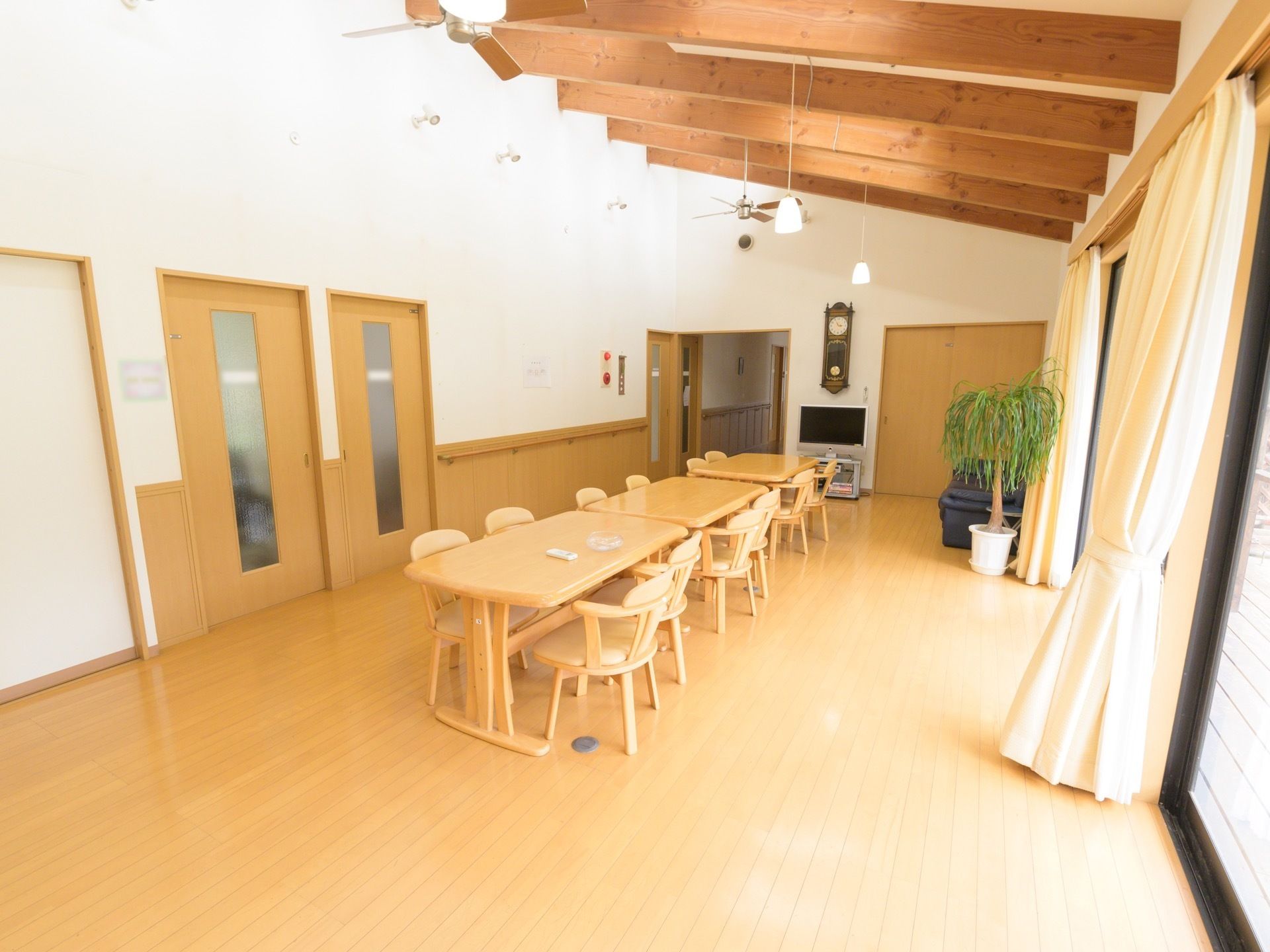 大きな窓から明るい自然光が差し込む開放的な食堂である。４人掛けダイニングテーブルが並べられており、テレビやソファが設置されている。