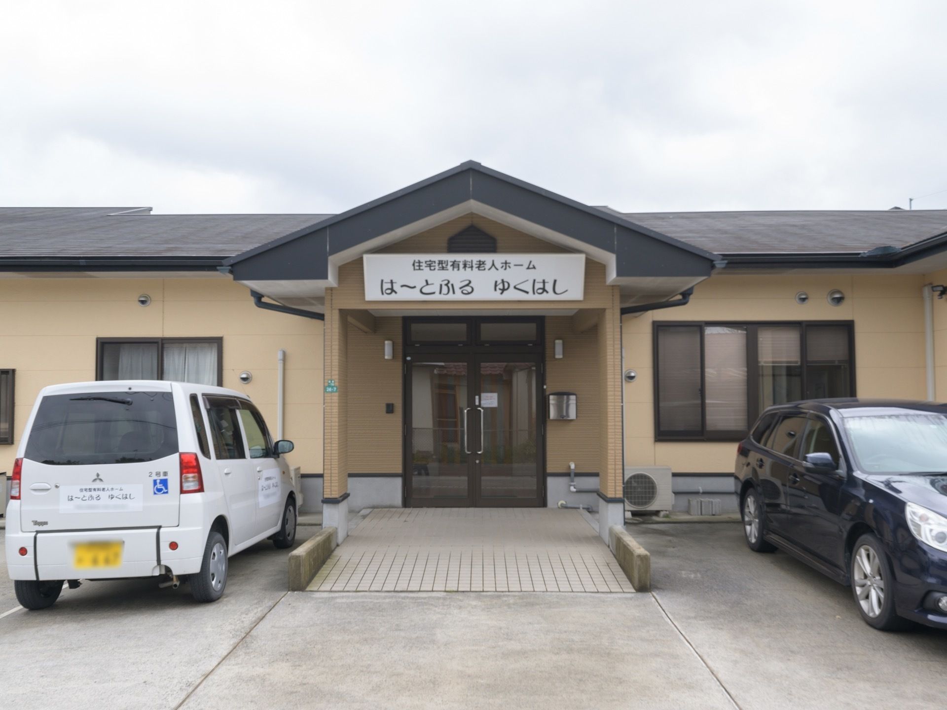 三角屋根の施設正面入口である。施設名称のプレートが掲げられ、両サイドには駐車スペースが設けられている。2台の車が停車している。