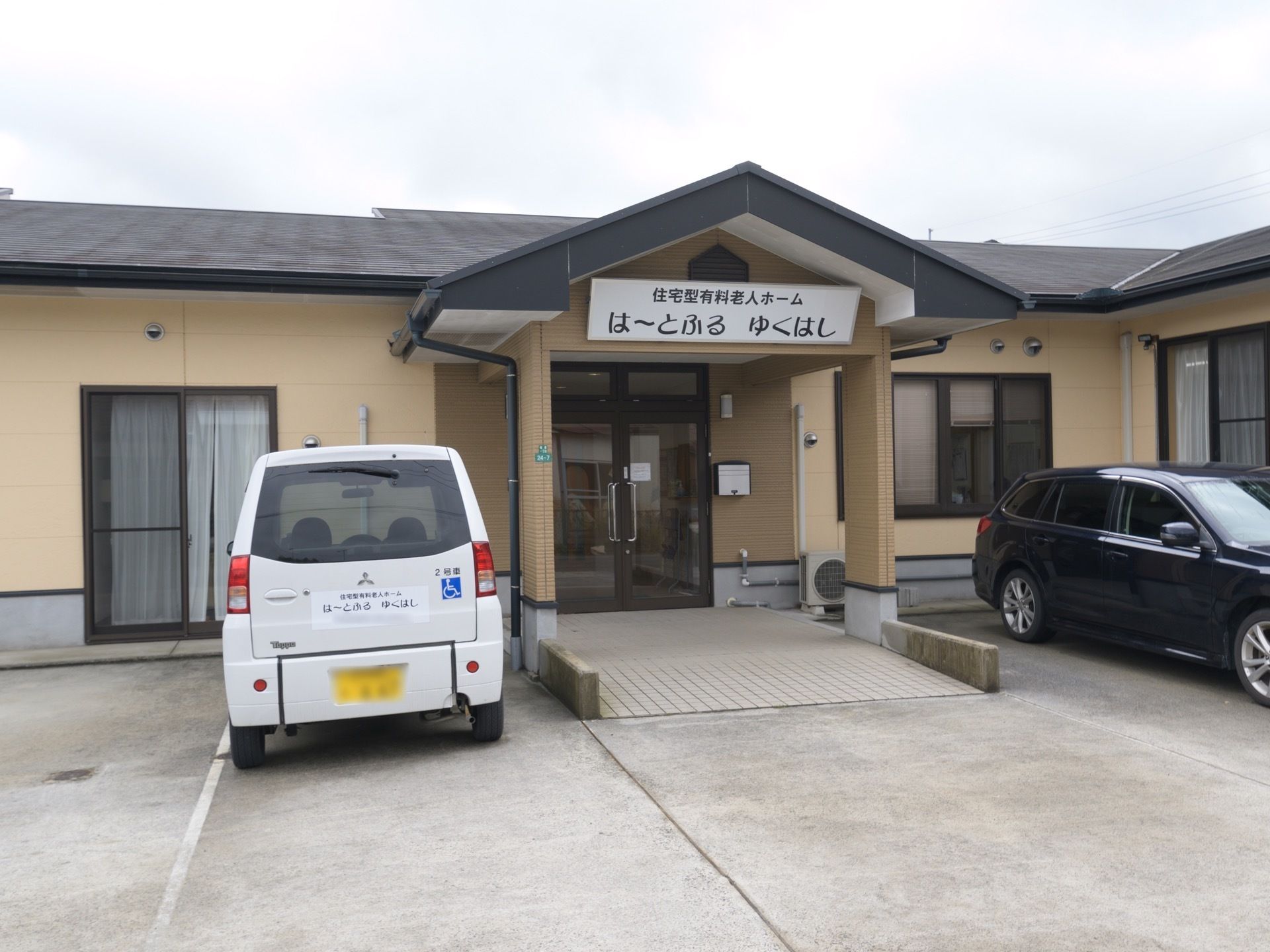 三角屋根が設置された入口前のポーチである。両サイドには駐車スペースが設けられ、乗用車と施設専用車が停車している。
