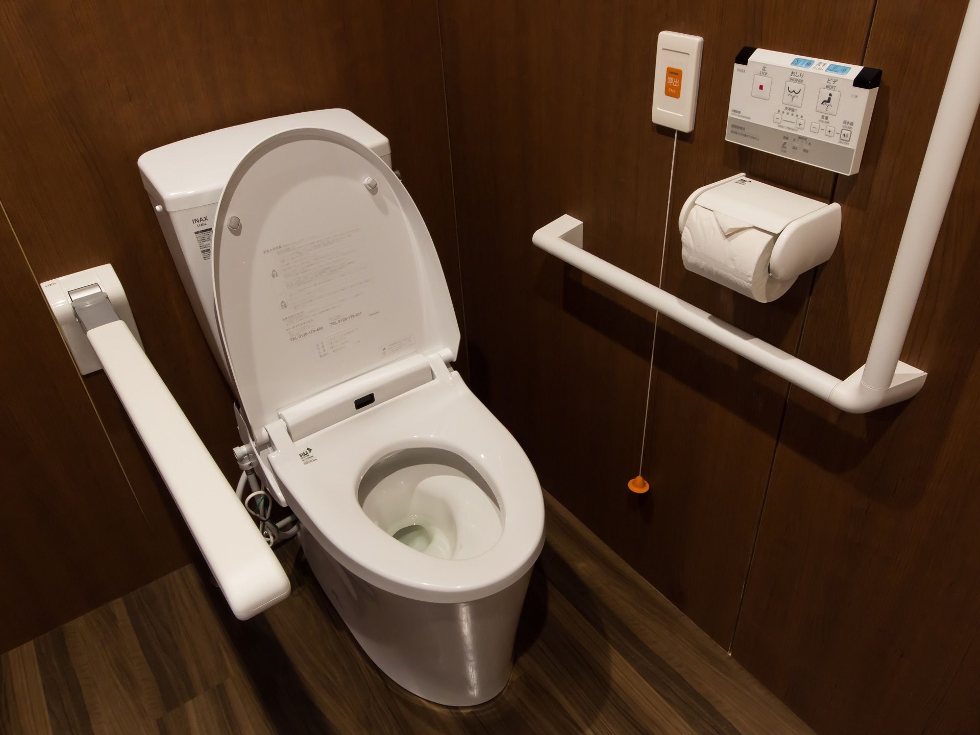 木目調の壁が落ち着いた雰囲気のトイレである。壁にはナースコールが設置されており、リモコンと手すりも取り付けられている。