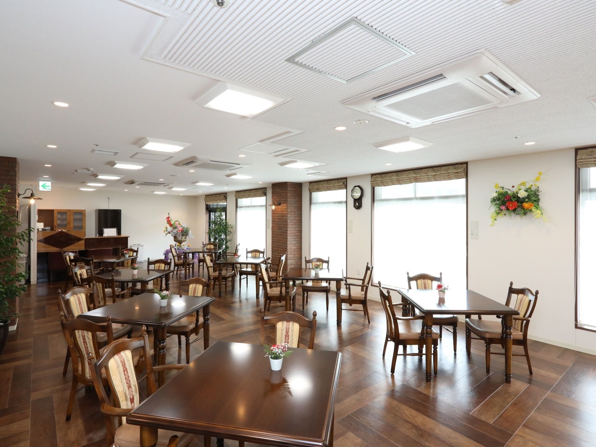 食堂に、茶色の天板のテーブルと、布張りの背もたれがついた椅子が多く置かれている。多くの照明が設置されている。