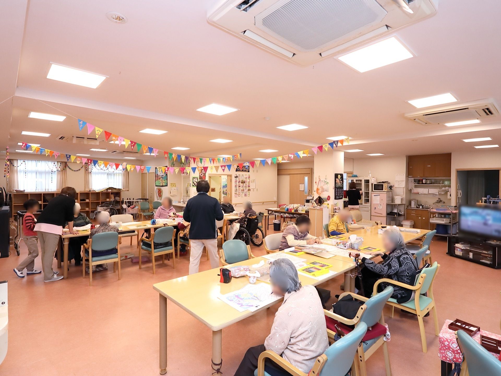 共有スペースに多くの入居者様が集まって、創作活動をしている。スタッフが２人いて、入居者様と話をしている。