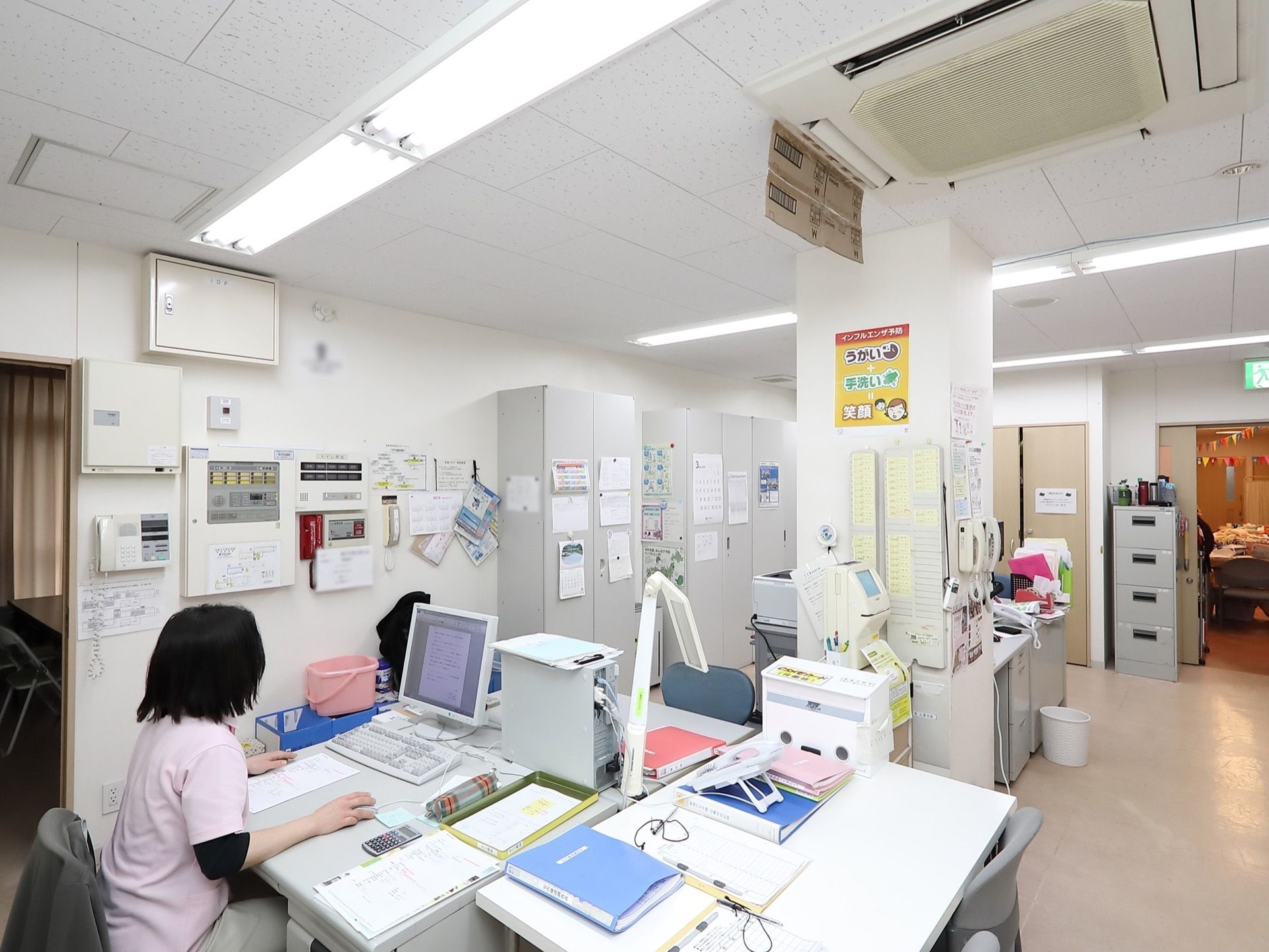 事務室で、女性スタッフがパソコンで作業をしている。壁際にはキャビネットが置かれ、その扉にはいろいろな掲示物が貼られている。