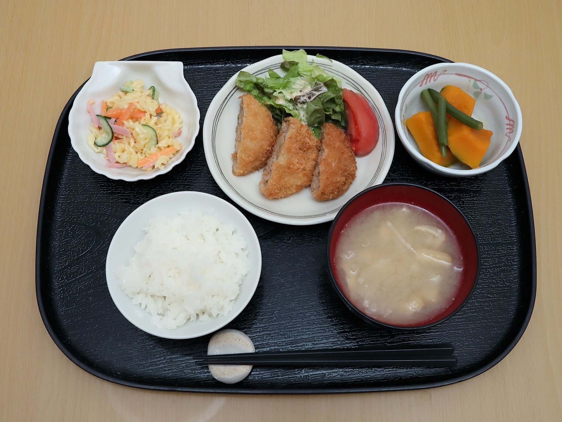 ご飯とみそ汁の他に、おかずが３皿、黒いお盆に載っている。手前には、箸置きがあり、黒色の箸が置かれている。