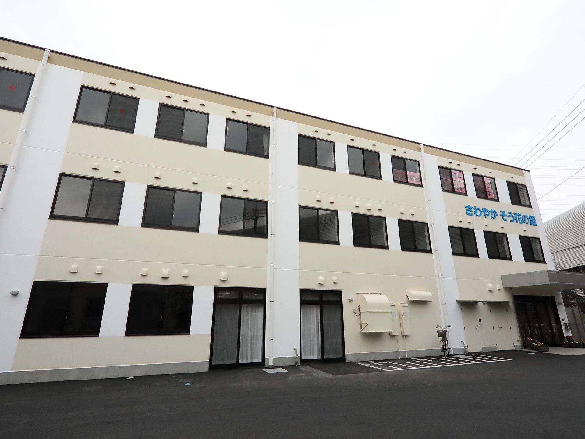 施設の建物は三階建てで、壁は明るいベージュ色である。前面は段差のないアスファルトで、玄関横に駐車スペースを設けている。