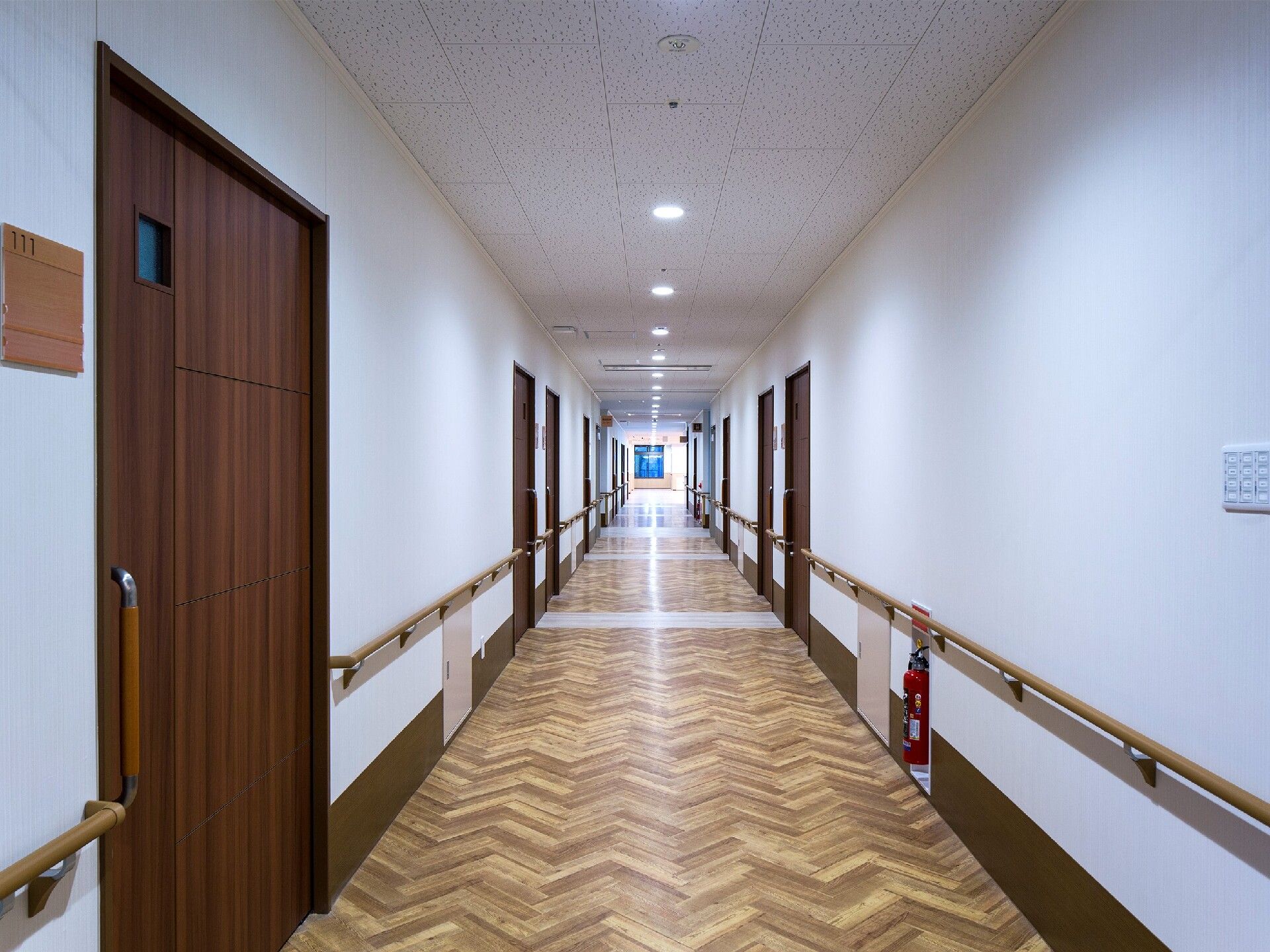 白を基調とした館内にはまっすぐな廊下があり、手すりと居室のドアが両サイドにあり、突き当りにはブルーの窓のようなものが見える。