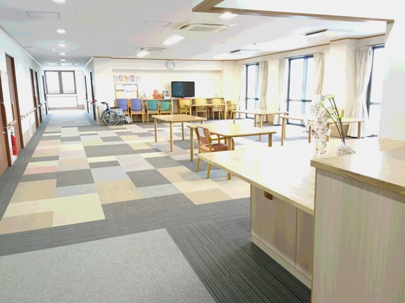 モザイク柄のグレーのカーペットが敷かれたリビングダイニングには木製のテーブルセット屋大きなテレビがあり、車いすも置かれている。