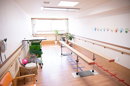白い花がたくさん咲いている道を楽しそうに散歩している車いすの高齢女性と付き添いの高齢男性。若い女性スタッフもそばにいる。