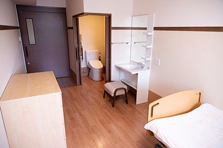 車いすの高齢女性が指さす方向を一緒に笑顔で見ているブルーのエプロンを付けた若い女性スタッフ。日当たりのいい室内で。