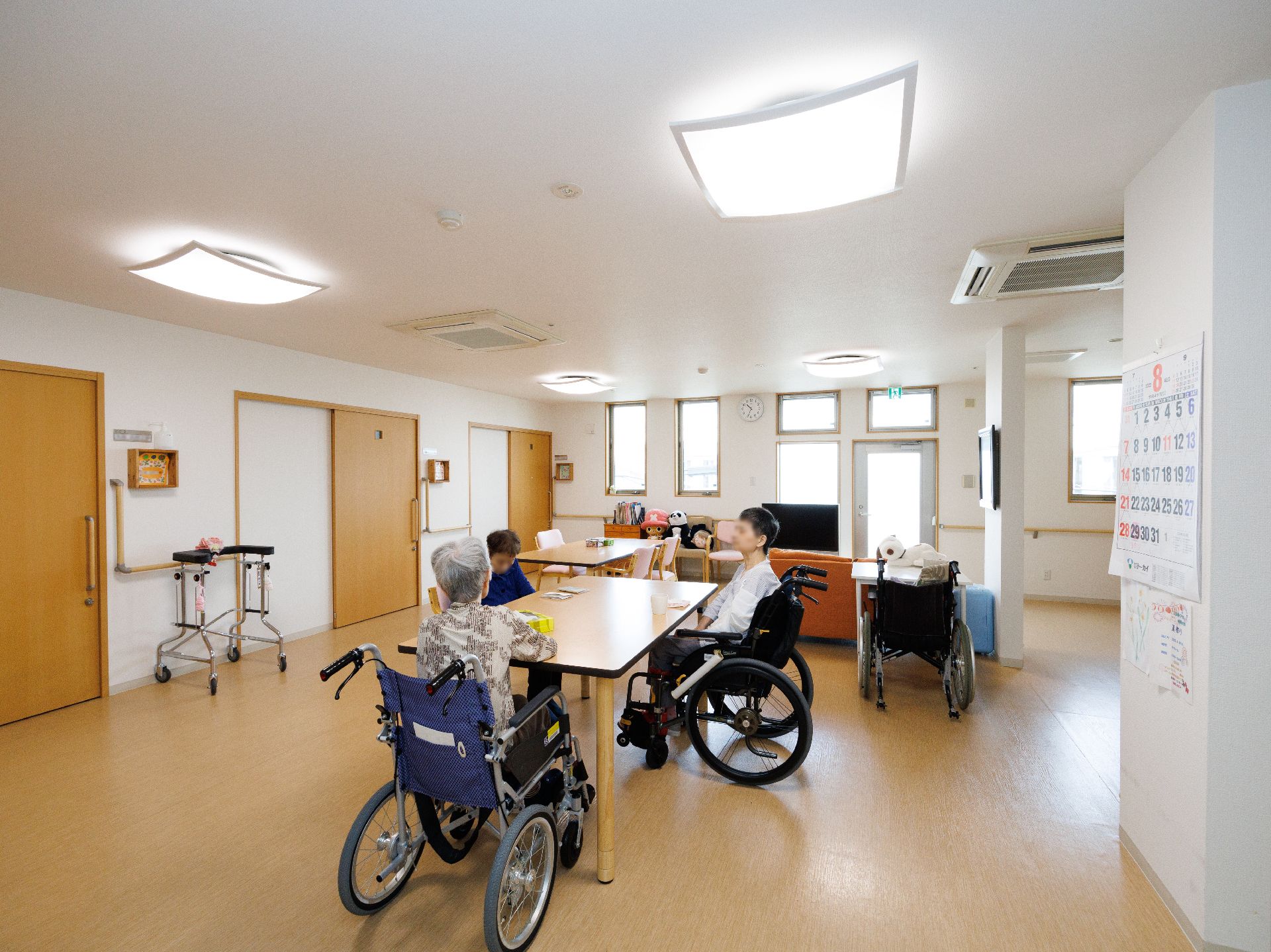 白い壁の広間に置かれた木製の机の周りに集まる入居者様