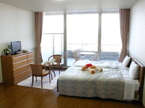 タンスやテーブルセット、寝具付きのベッドが２台設置された居室。窓の外にはベランダもあり、日当たりもよい。