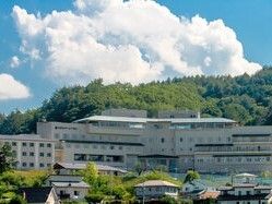 5階建ての建物はベージュで窓が多く、山の中腹にある。その下には住宅が建っている場所もあり、周辺には緑が多い。