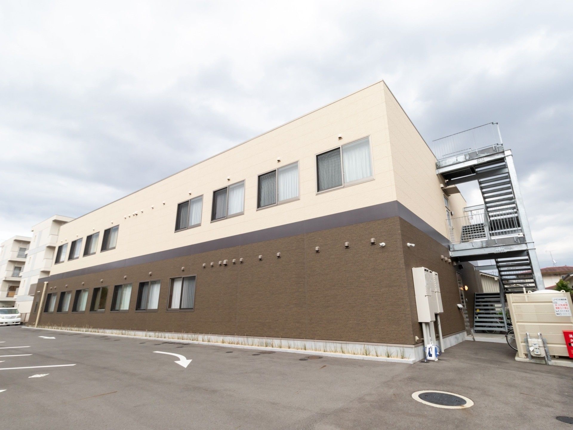 アスファルト舗装された敷地に、２階建て住宅が建っている。建物の横には非常用の外階段が設置されている。