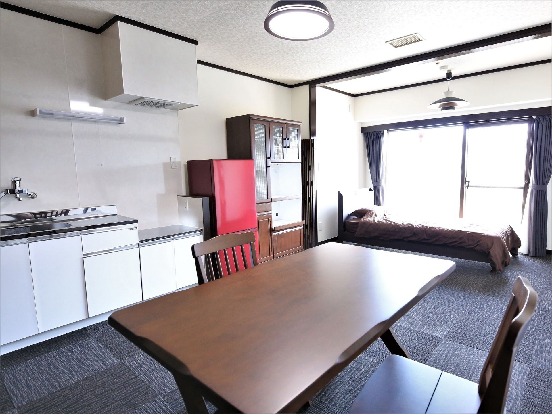 大きな窓が特徴的な居室。白く広々としたキッチンが備え付けられており、大きなテーブルやベッドが置かれている。