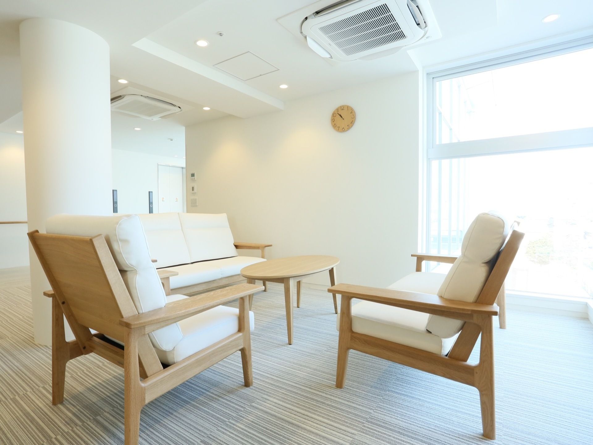 白い壁と大きな窓がある明るい空間に、丸いテーブルとオシャレなソファが置かれている。壁には時計が掛かっている。