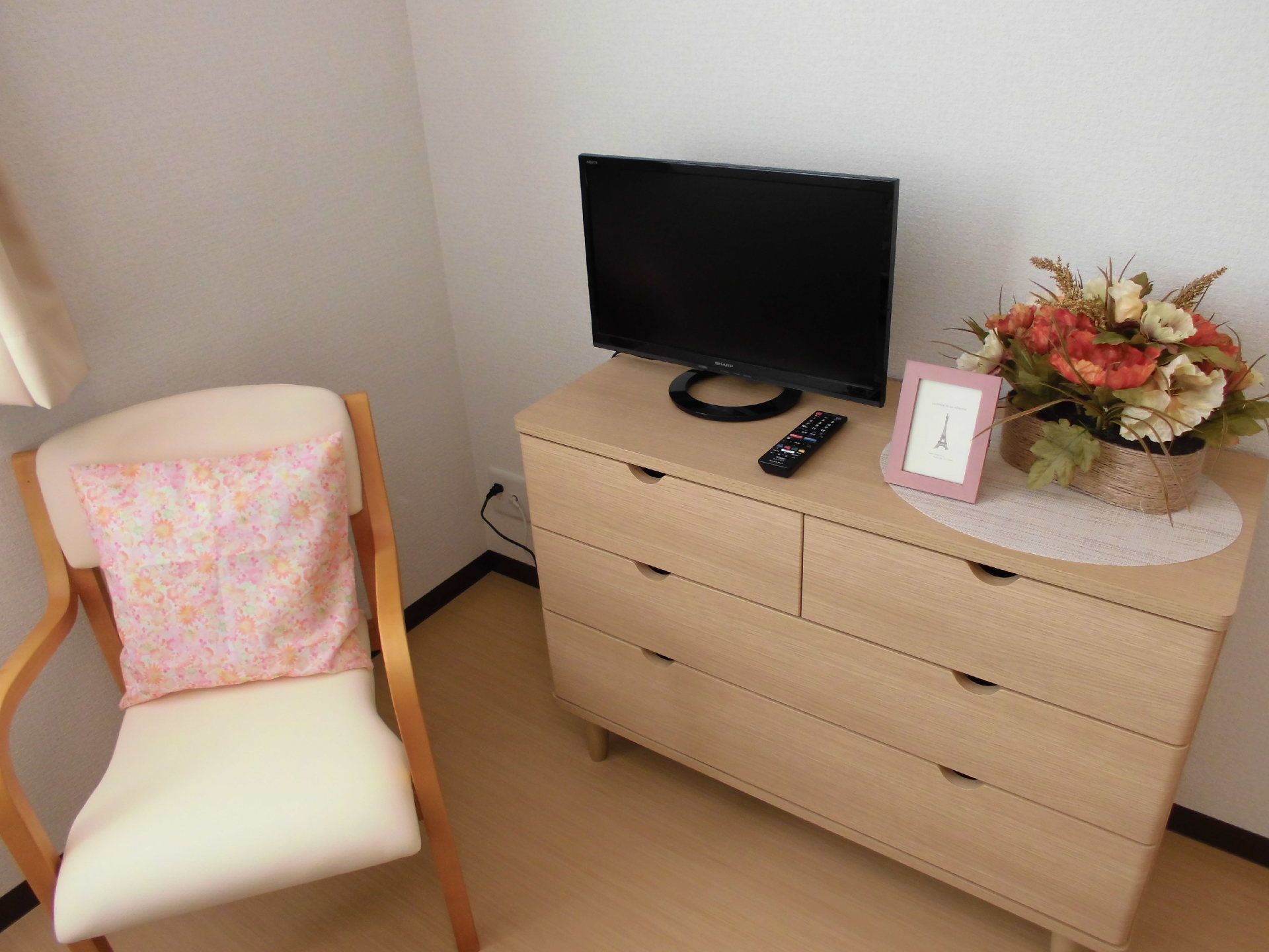 白いいすに3段のチェスト、その上には花やテレビが飾られている。