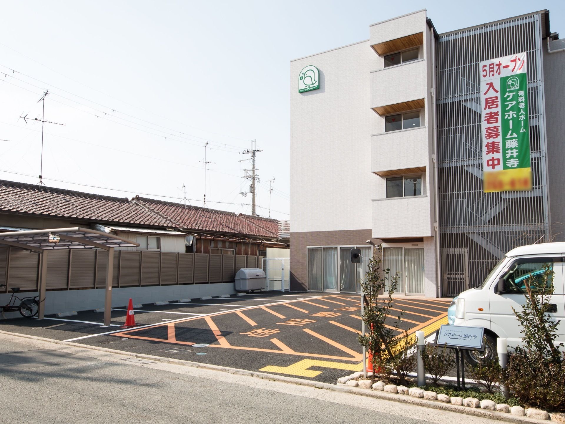 前の道路から見た施設の様子。敷地はアスファルト舗装の駐車場になっており、右に仕切り代わりの植え込みがある。