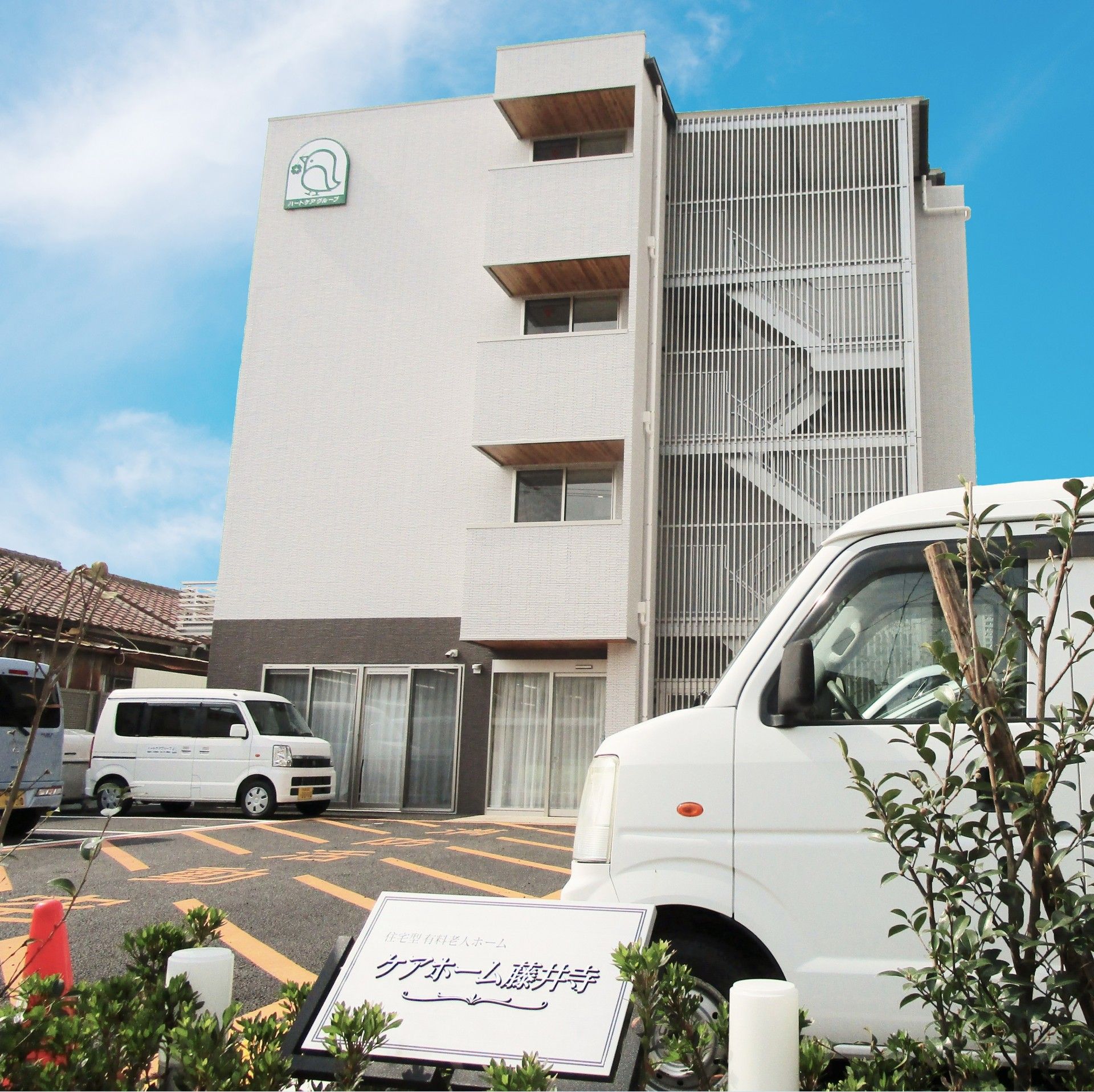 前の道路から見た施設の様子。敷地はアスファルト舗装の駐車場になっており、右に仕切り代わりの植え込みがある。