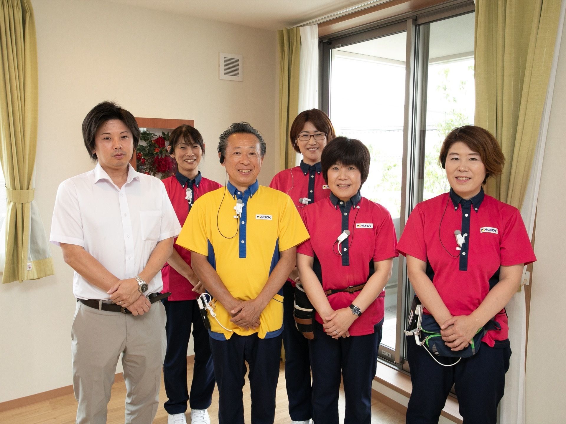 施設のスタッフが後ろに2人、前に4人笑顔で並んでいる。女性は赤色、男性は黄色のポロシャツを着用している。