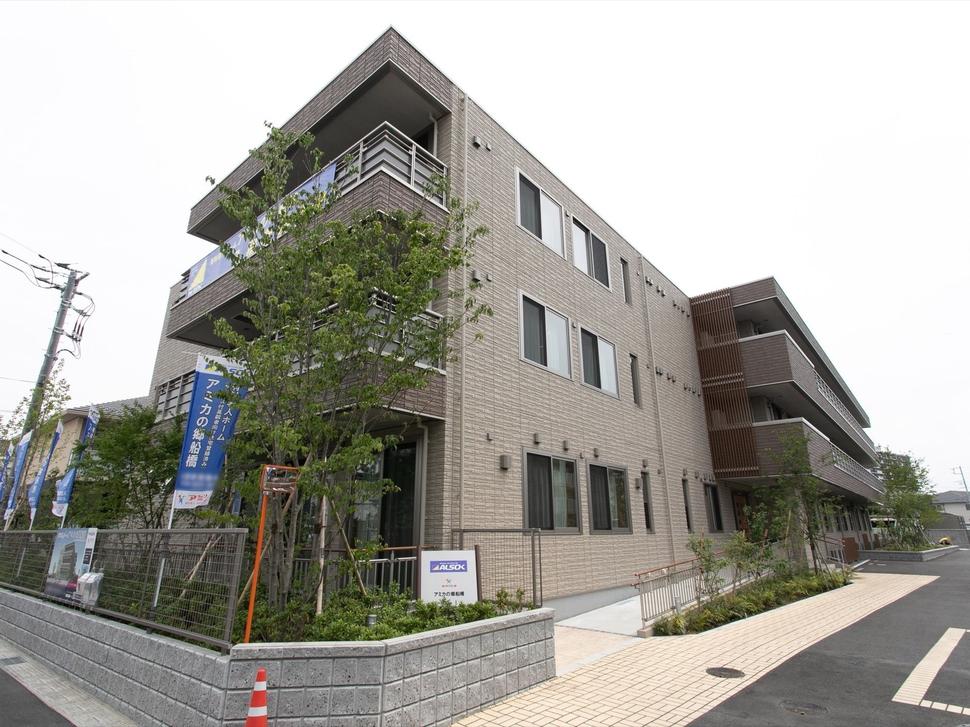 施設は3階建てで、グレーの外観となっている。周りにはたくさんの植物や木が植えられていて、玄関前にはスロープもある。