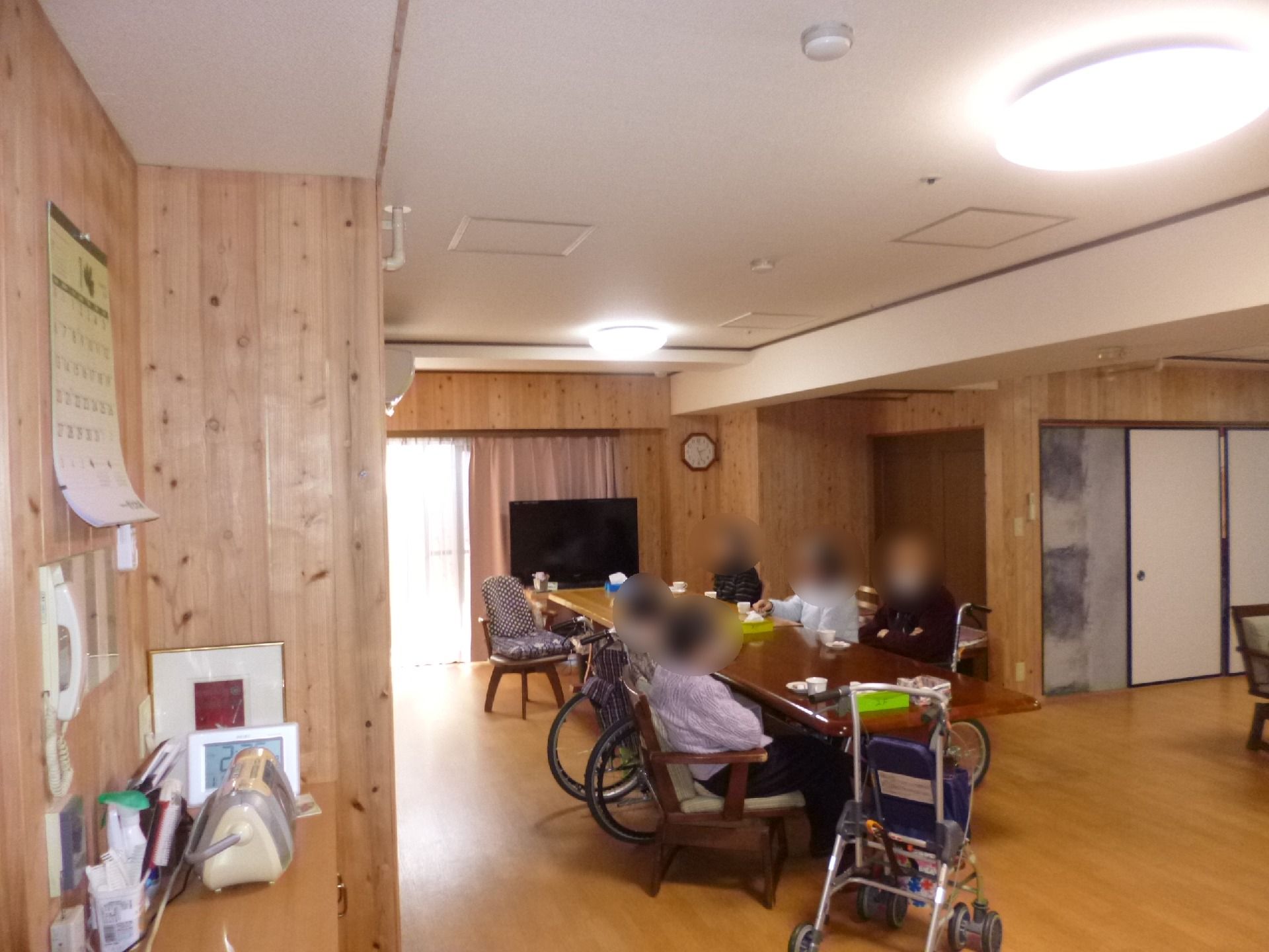 木製の机がおかれた部屋。　壁は木でできている。机には高齢者が何人か座っている