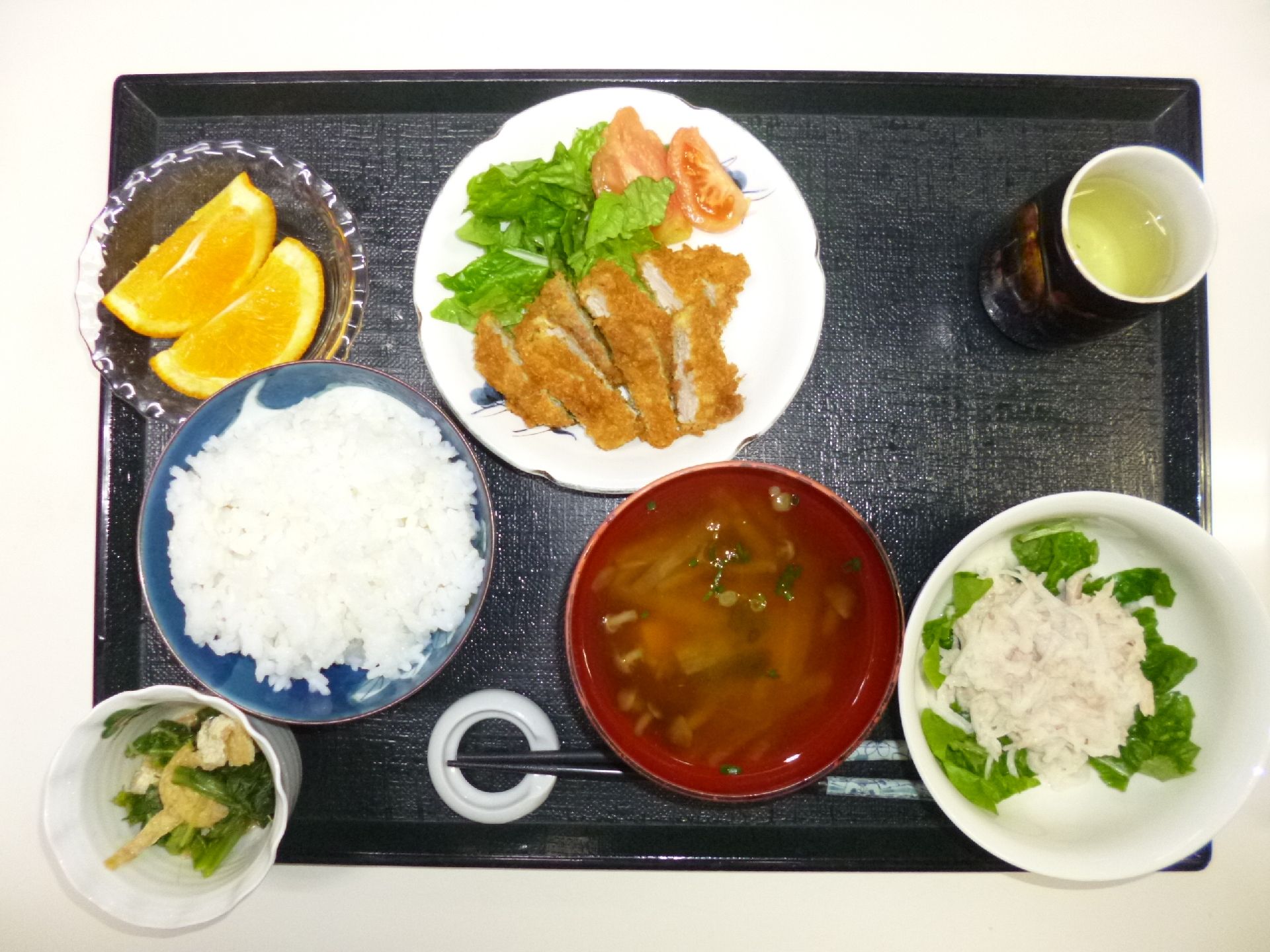黒いお盆の上にカツ、フルーツ、ご飯、お味噌汁が乗っている
