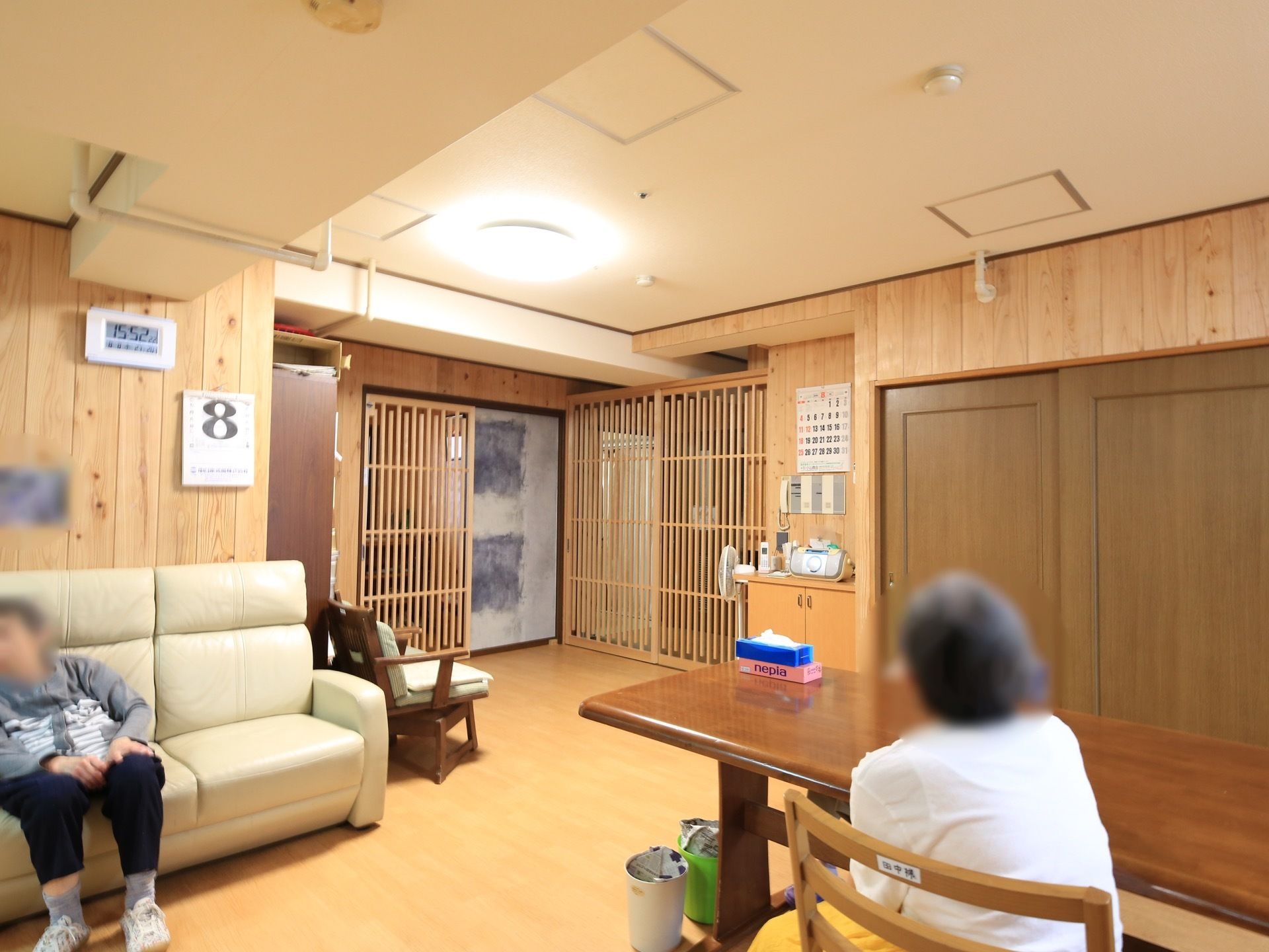 入居者様がソファーや椅子に座って寛いでいる。木目調の壁には、カレンダーやデジタル時計が掛けられている。
