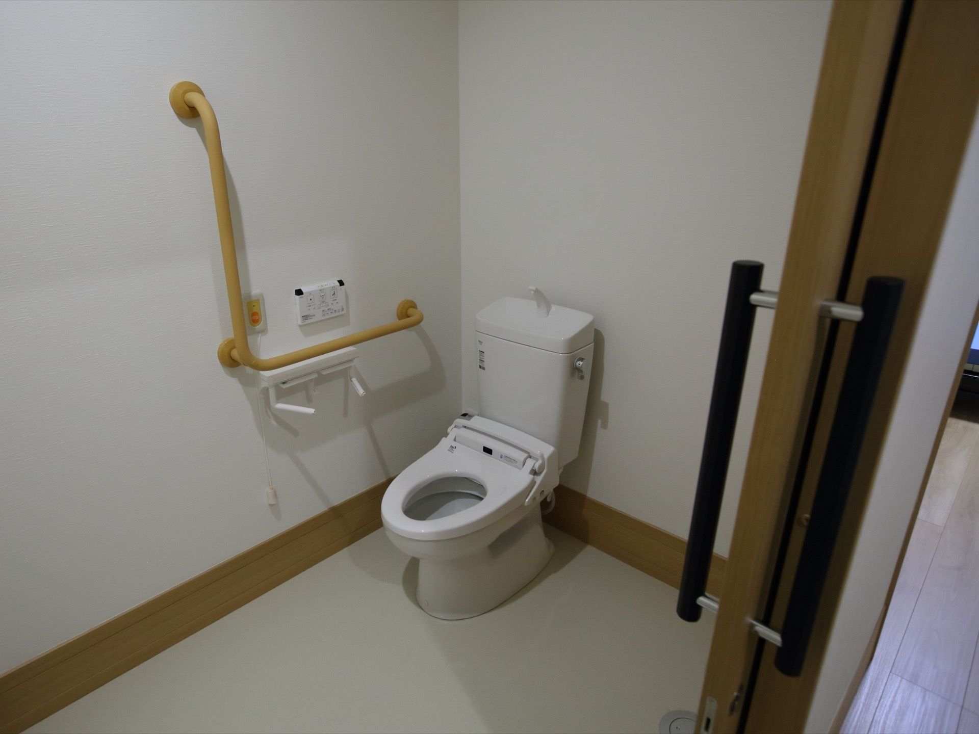 白色を基調としたトイレである。洋式の便座が備えられている。壁側には、Ｌ型の手すりがある。