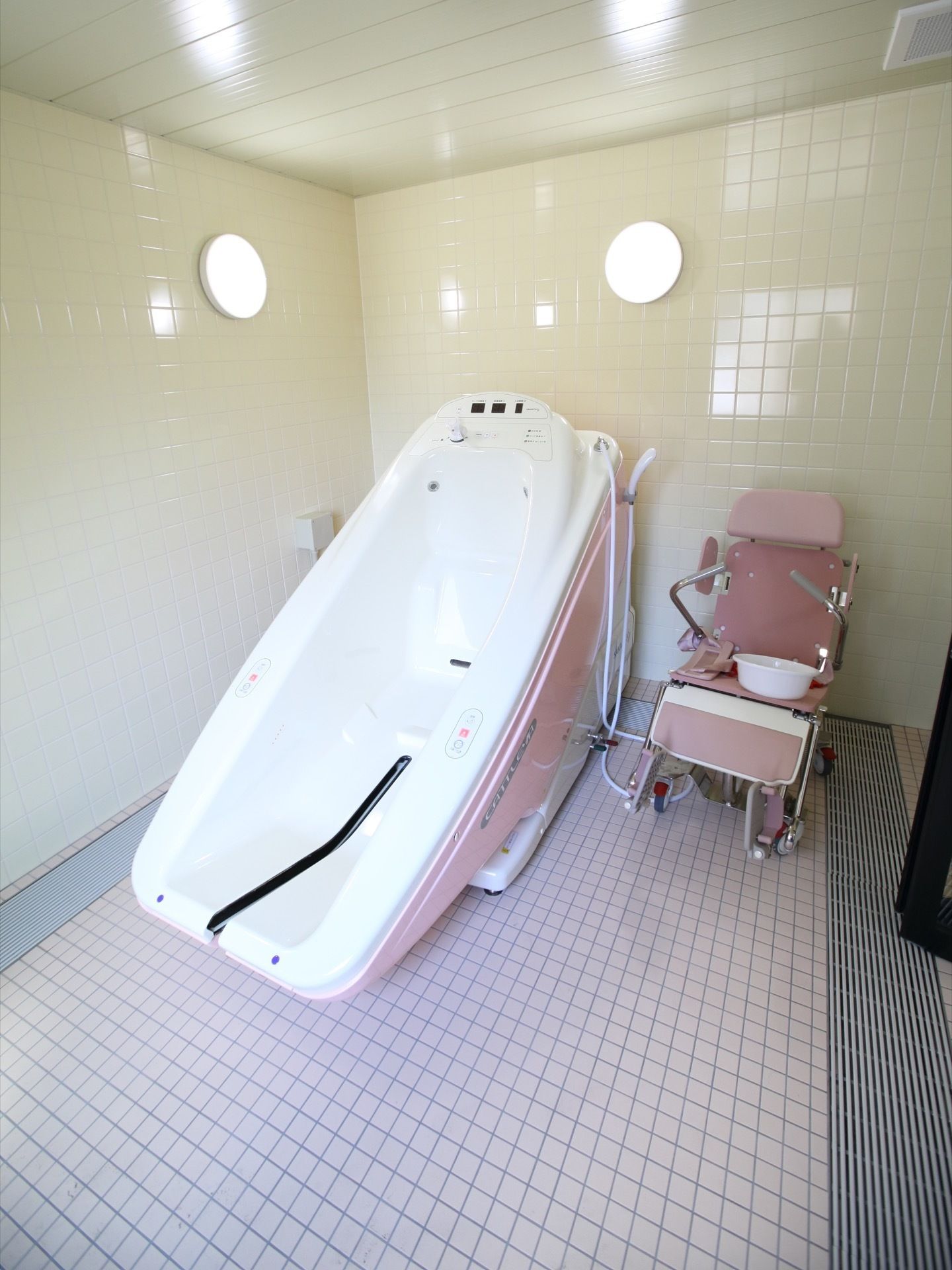 白色の壁、淡いピンク色のタイル床が設置された機械浴室である。広々とした介護専用の浴槽が置かれている。