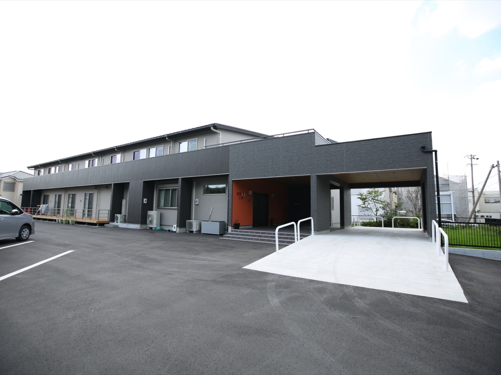 落ち着いた色調の２階建て施設である。居室にバルコニーがある。白線が引かれた、駐車場も完備してある。