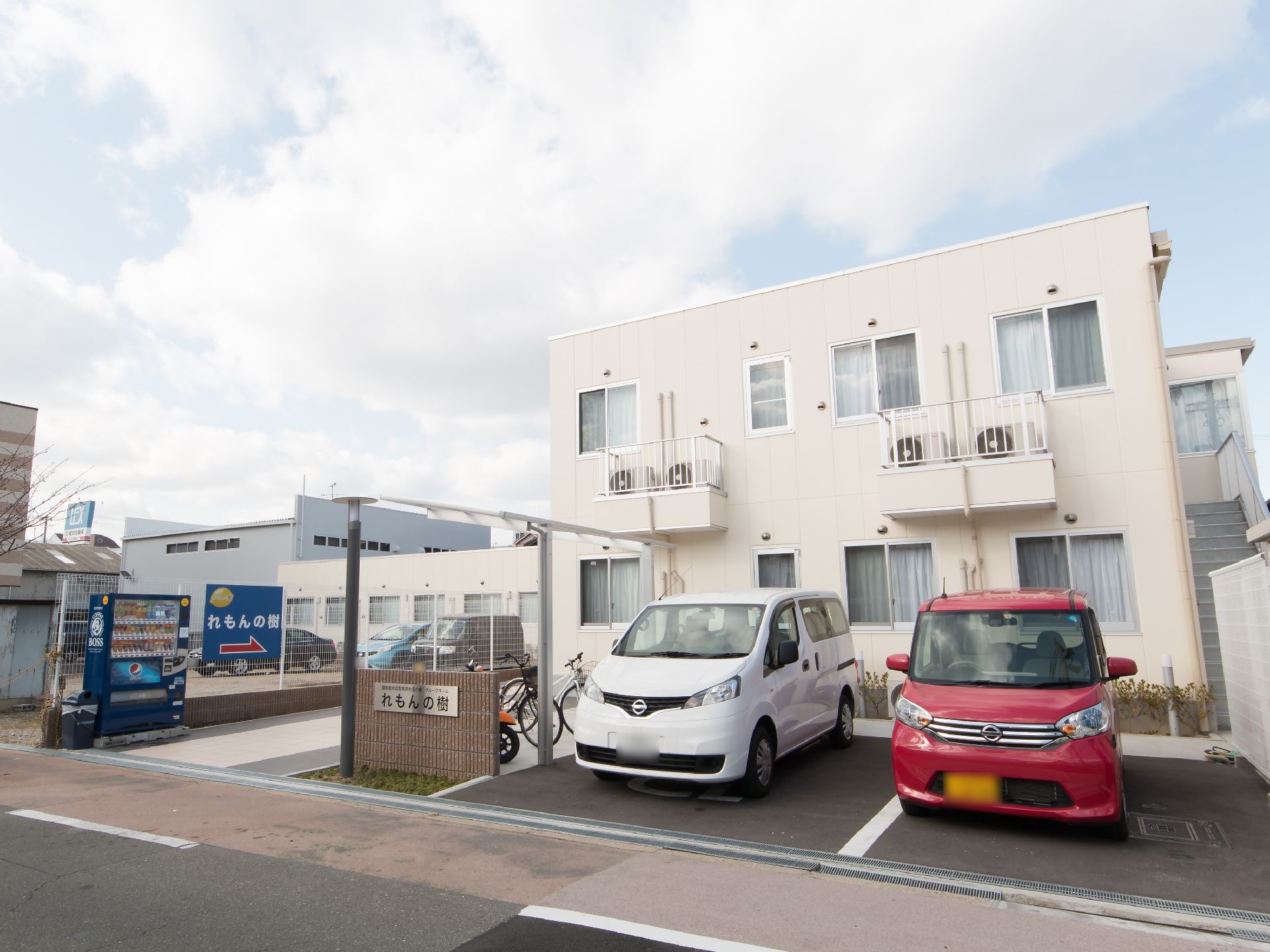 施設の前に駐車場と駐輪スペースが設けられている。駐輪スペースには屋根が設置されている。施設名が書かれた看板がフェンスについている。