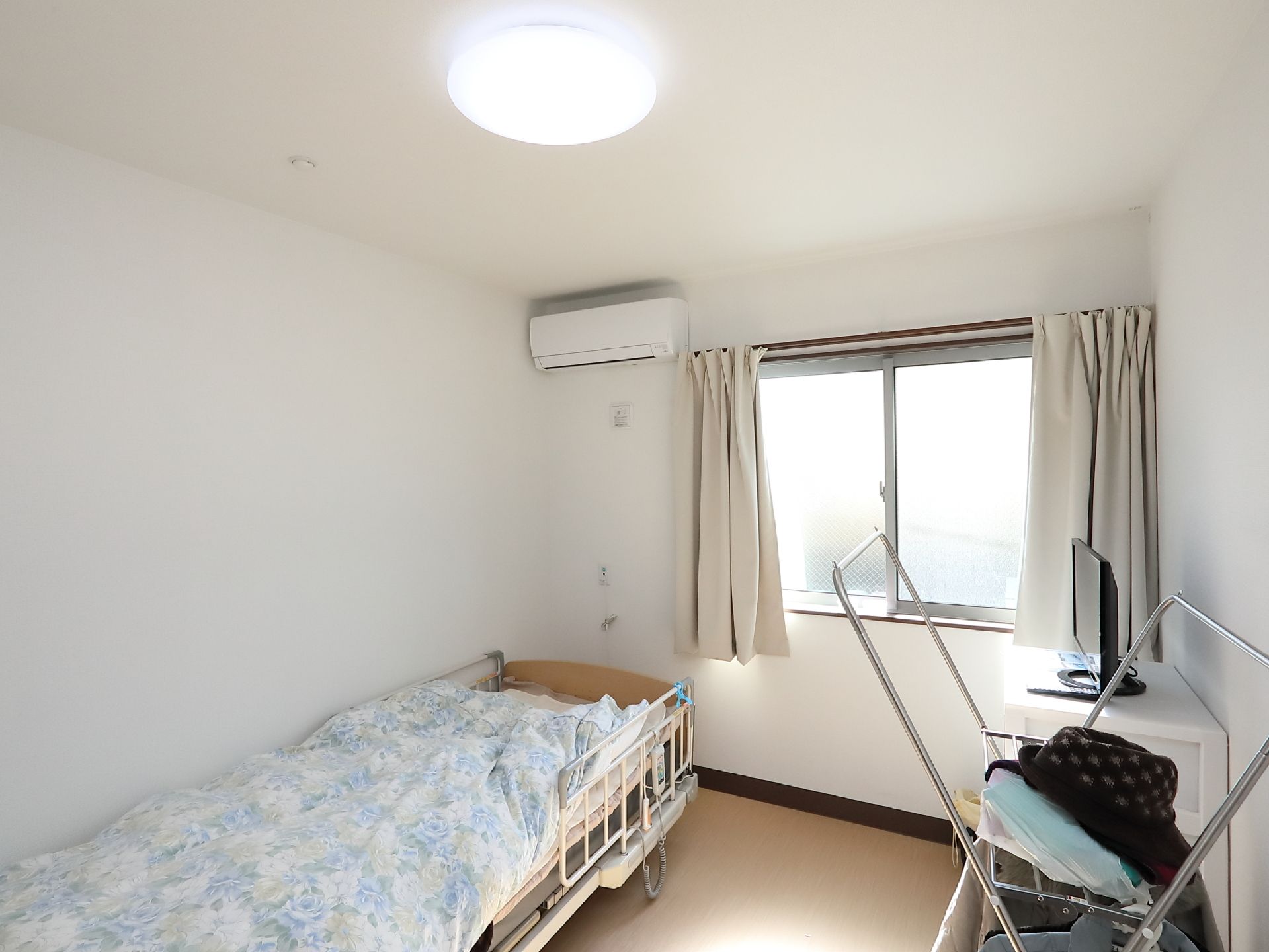 居室には転落防止柵つきのベッドやエアコンが設置されている。収納ケースの上に小さな画面のテレビが置かれている。