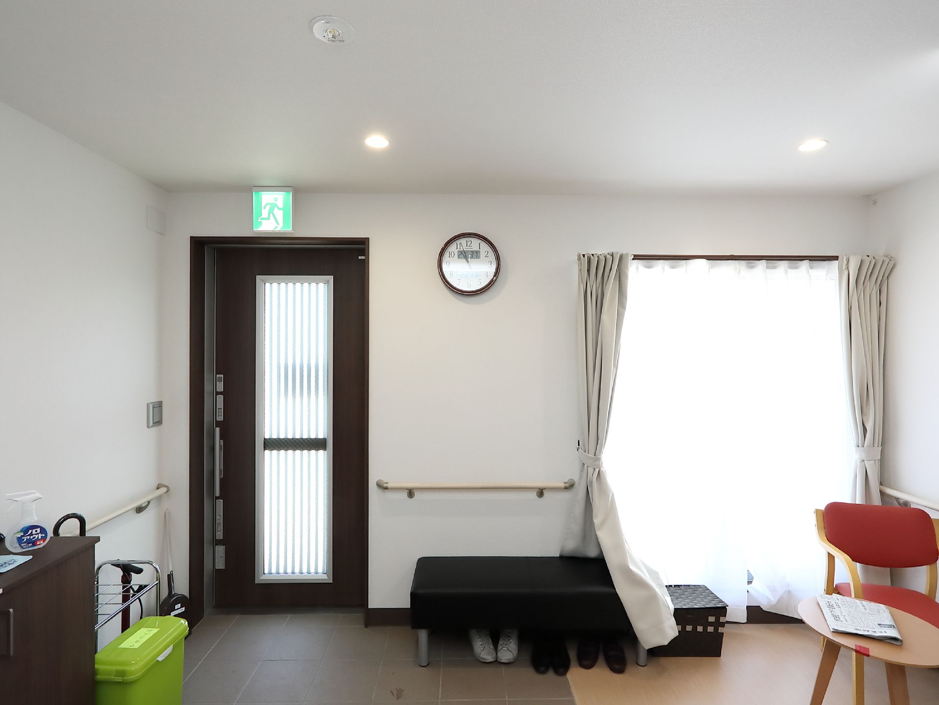 玄関はすりガラスが入った黒色の引き戸になっている。扉の上には、非常口のマークがある。黒色のベンチの下に靴が３足置かれている。