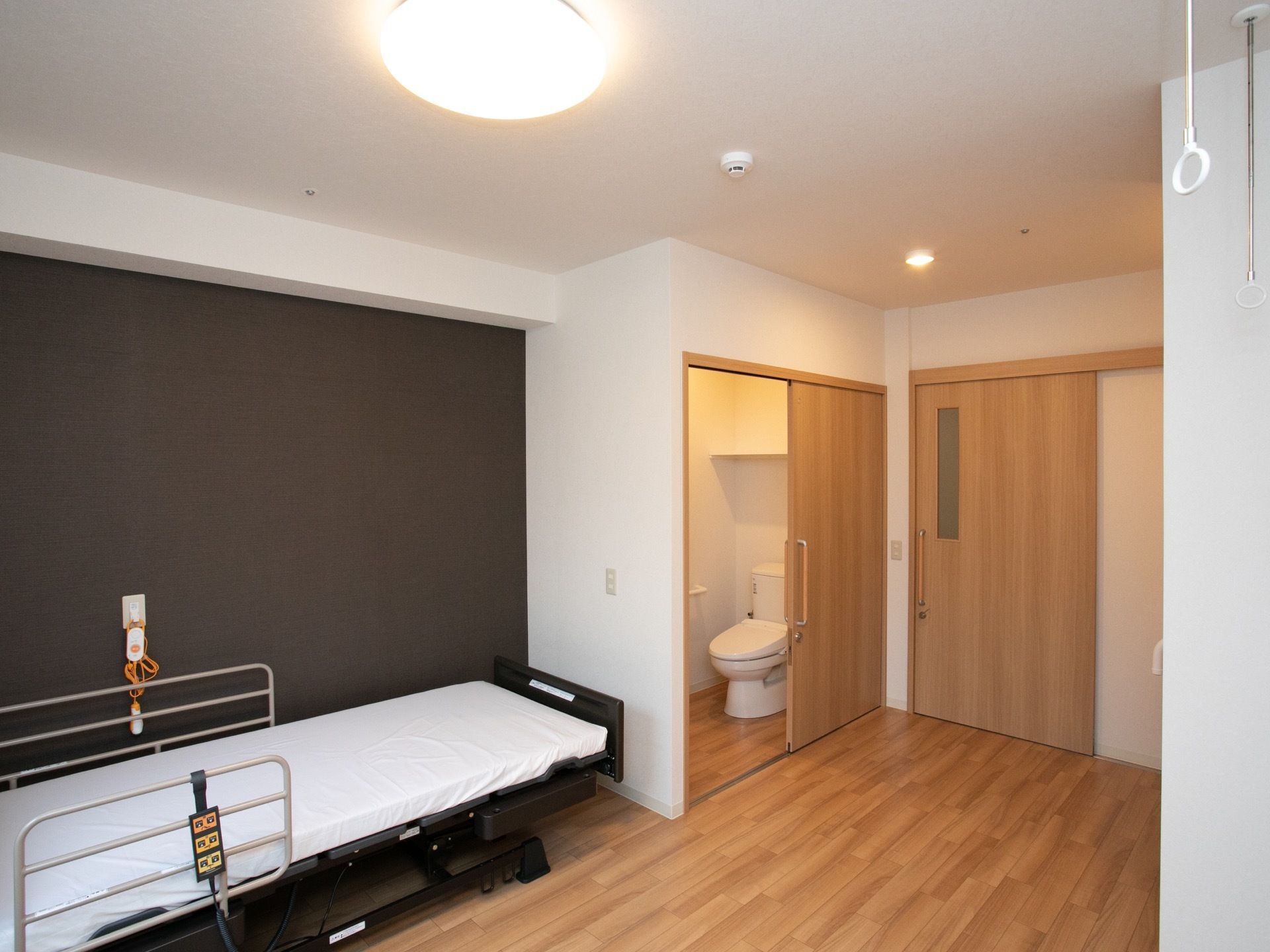 居室のドアは、ライトブラウンの大きな引き戸である。そのすぐ左側にトイレがある。丸い形の照明器具が、天井に付いている。