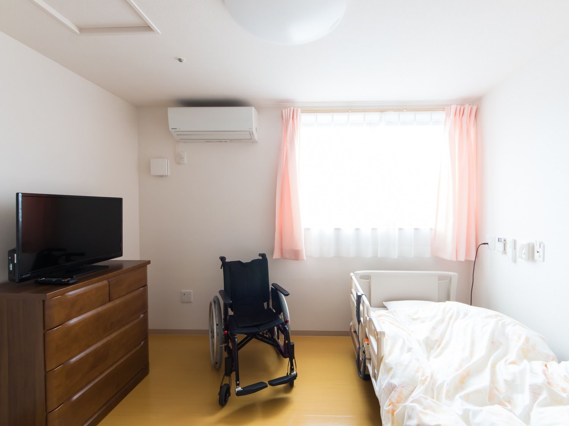 居室に電動ベッドと車椅子、テレビ、タンスが置かれている。突き当たりには腰の高さに窓があり、窓の横にはエアコンがある。