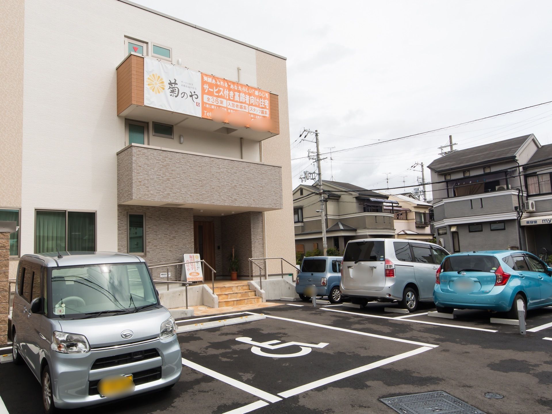 ３階建てのマンションタイプの建物。前が駐車スペースになっていて、車が4台停まっている。周りには戸建て住宅が多い。
