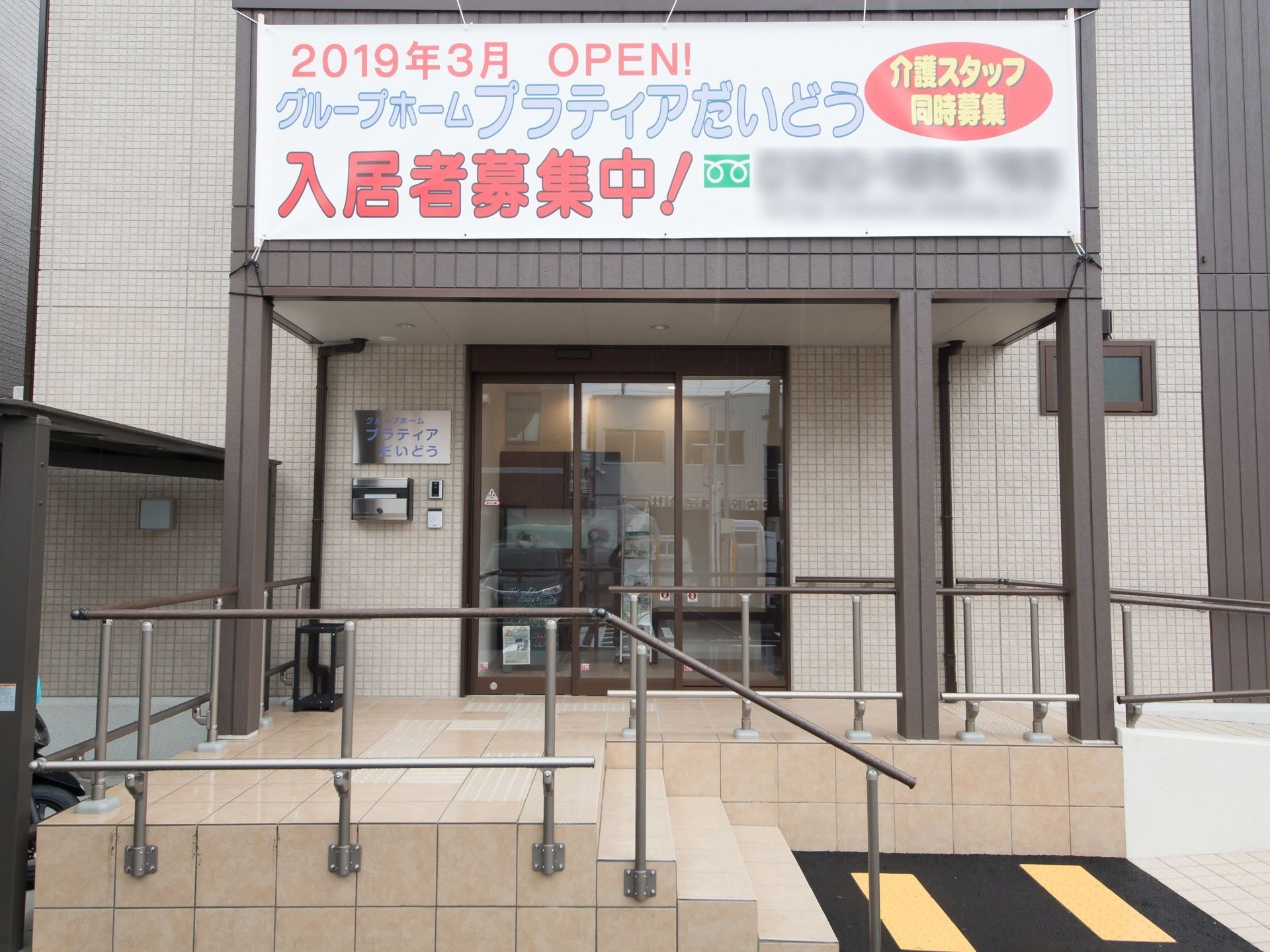施設の玄関へのアプローチには階段とスロープが設置されている。外壁はベージュを基調としている。扉はガラス張りになっている。
