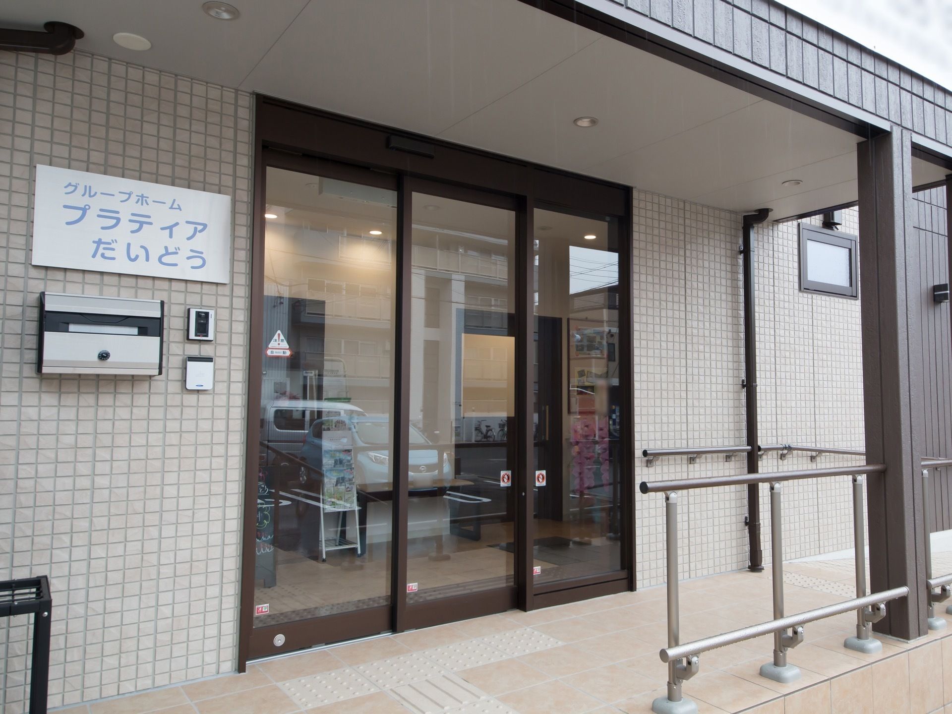 施設の入り口の外壁はベージュを基調としている。扉は広く開閉する３枚タイプになっている。玄関前に屋根が設置されている。