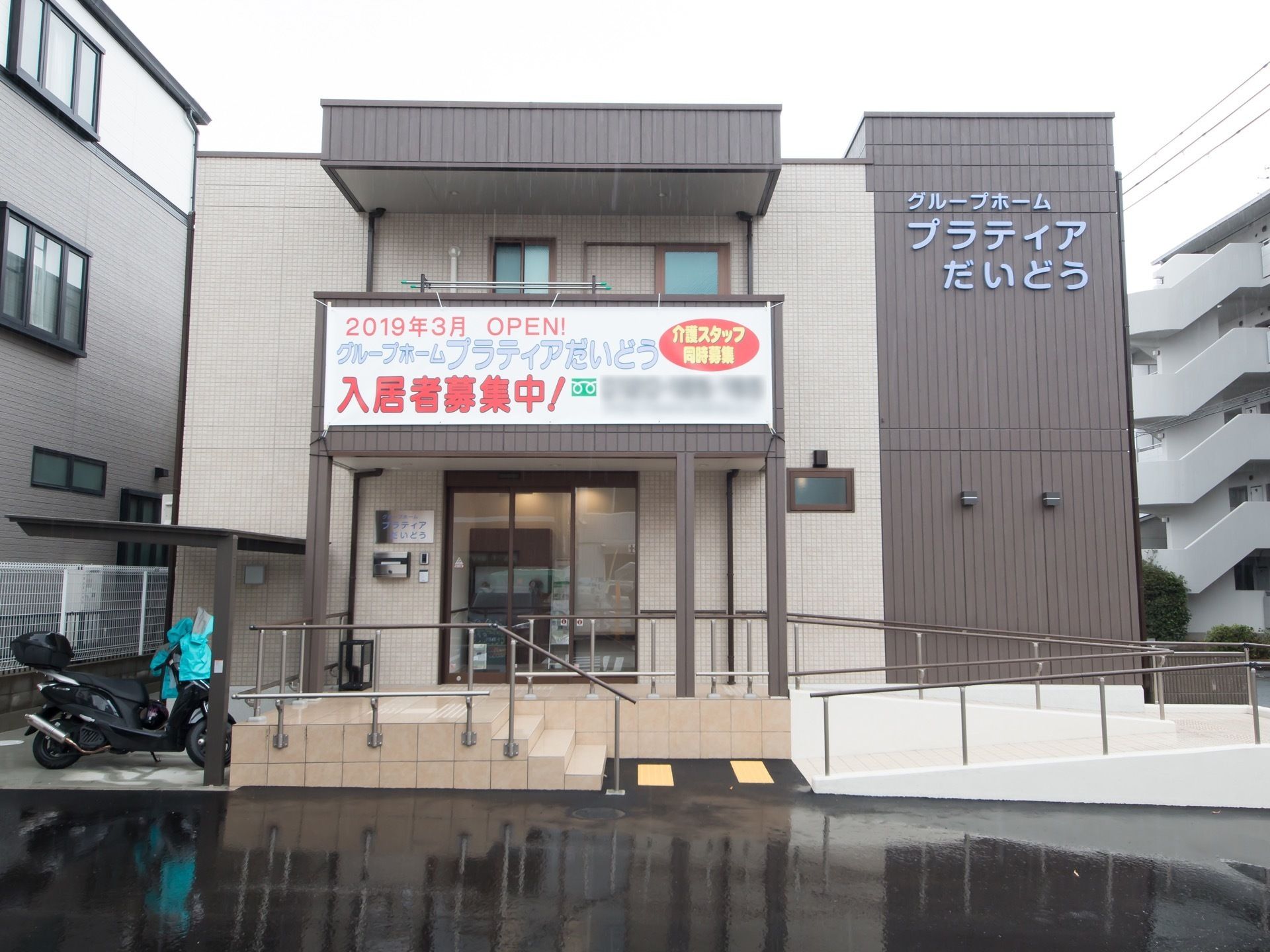 ベージュとダークブラウンのツートンカラーの施設建物である。玄関周りに、階段とスロープ、手すりを設けている。