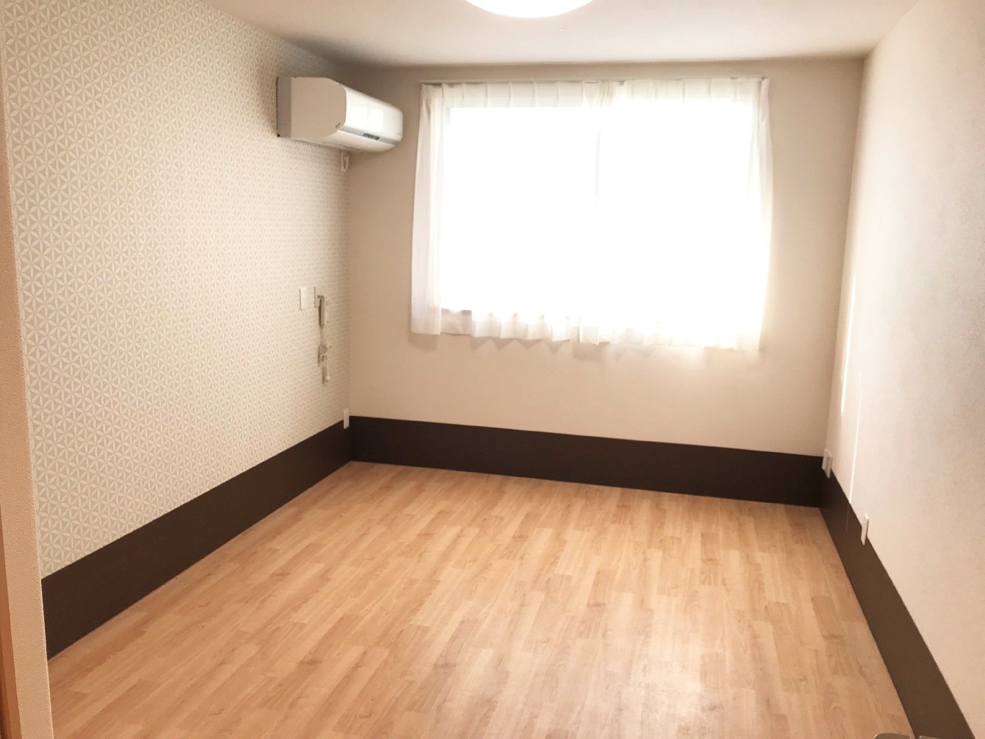 フローリング貼りの床の居室。窓にはレースが掛かっており、壁にはエアコンが設置されている。