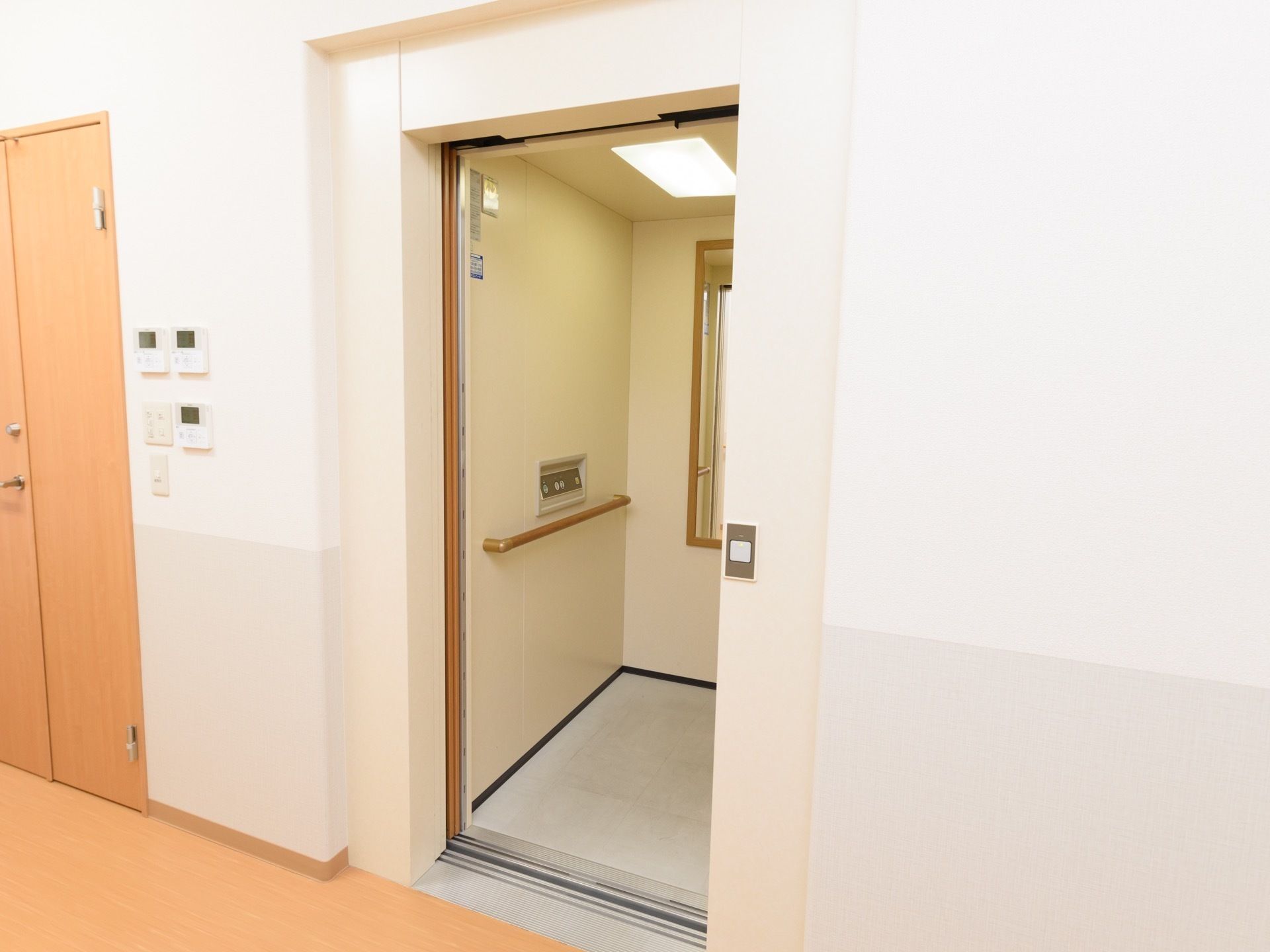 白と淡いグレーの壁の廊下に、エレベーターが設置されている。内部には鏡と、木製の手すりが設置されている。