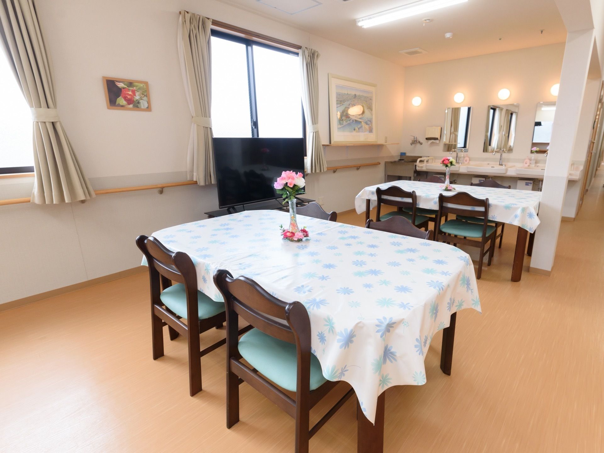 テーブルクロスがかけられた４人掛けのテーブルが２台並んでおり、それぞれに花瓶に入った花が飾られている。テレビや洗面台が設置されている。