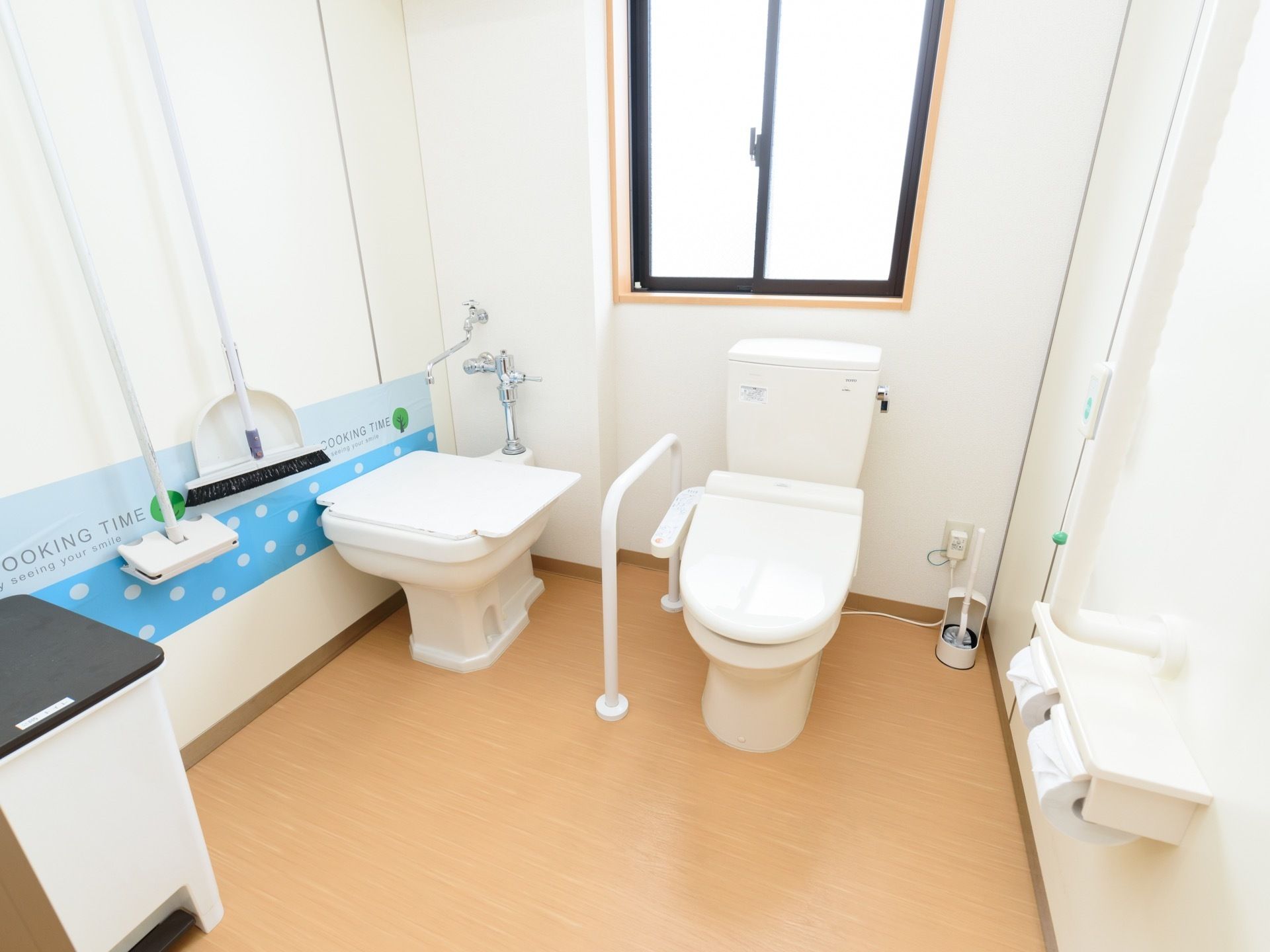 広い個室内に、２種類の便器が備わっている。壁には掃除用の箒やちりとりが掛けられ、手すりやナースコールが設置されている。